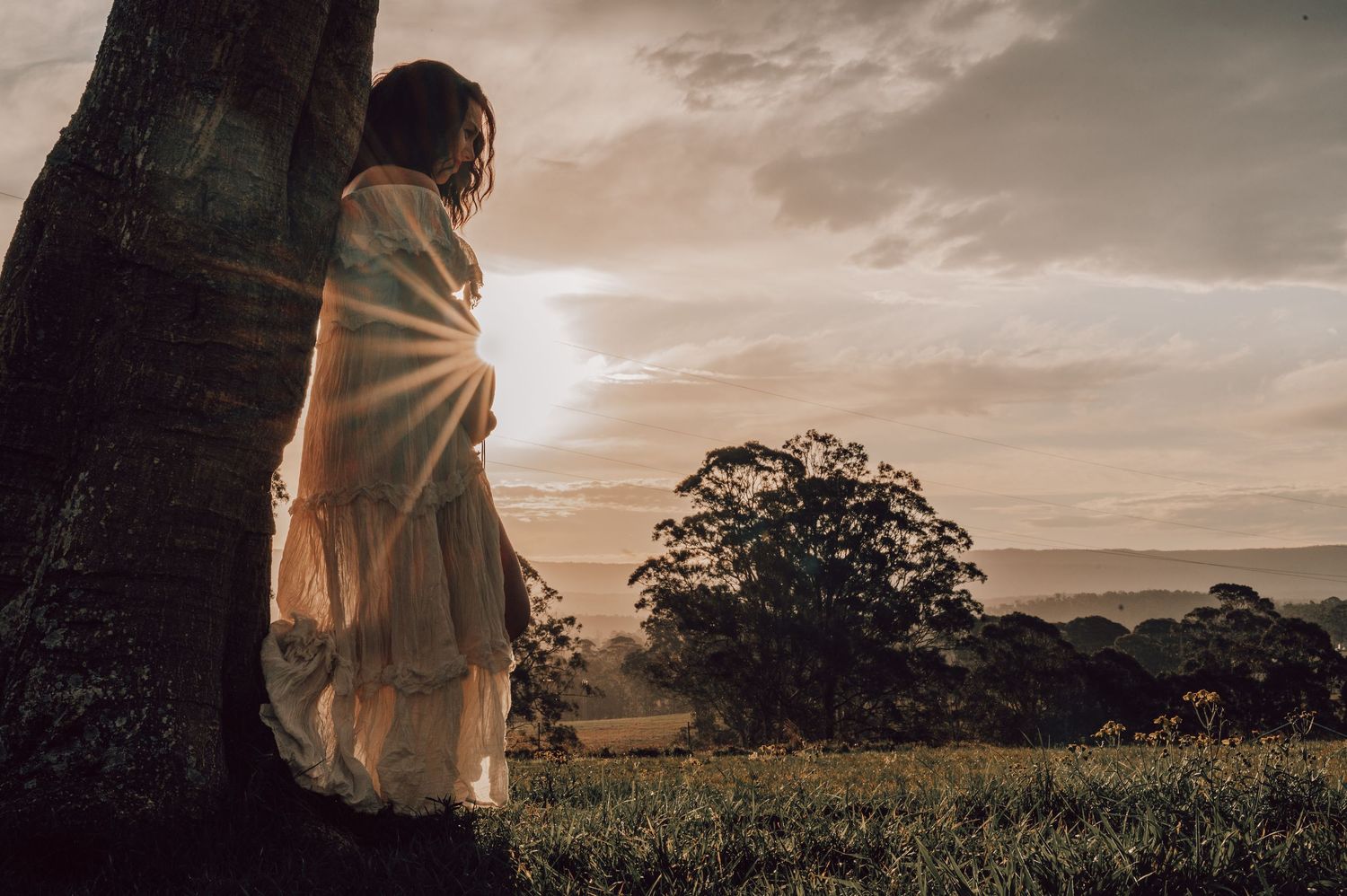Woman in flowing white dress leaning against tree watching golden sunset over valley.