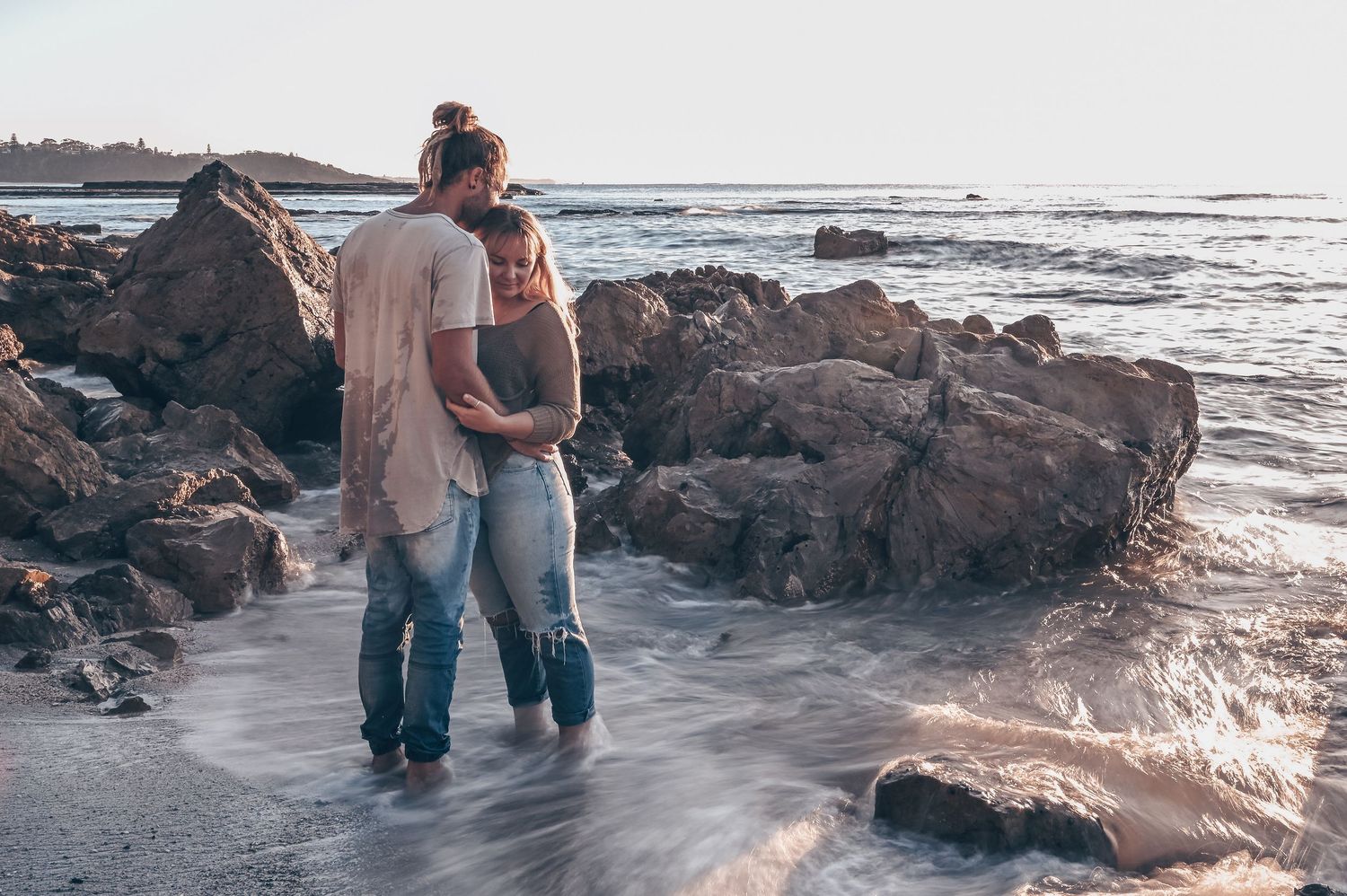 Couple embracing while standing in ocean waves among rocky coastal shoreline at sunset. Couple Photography