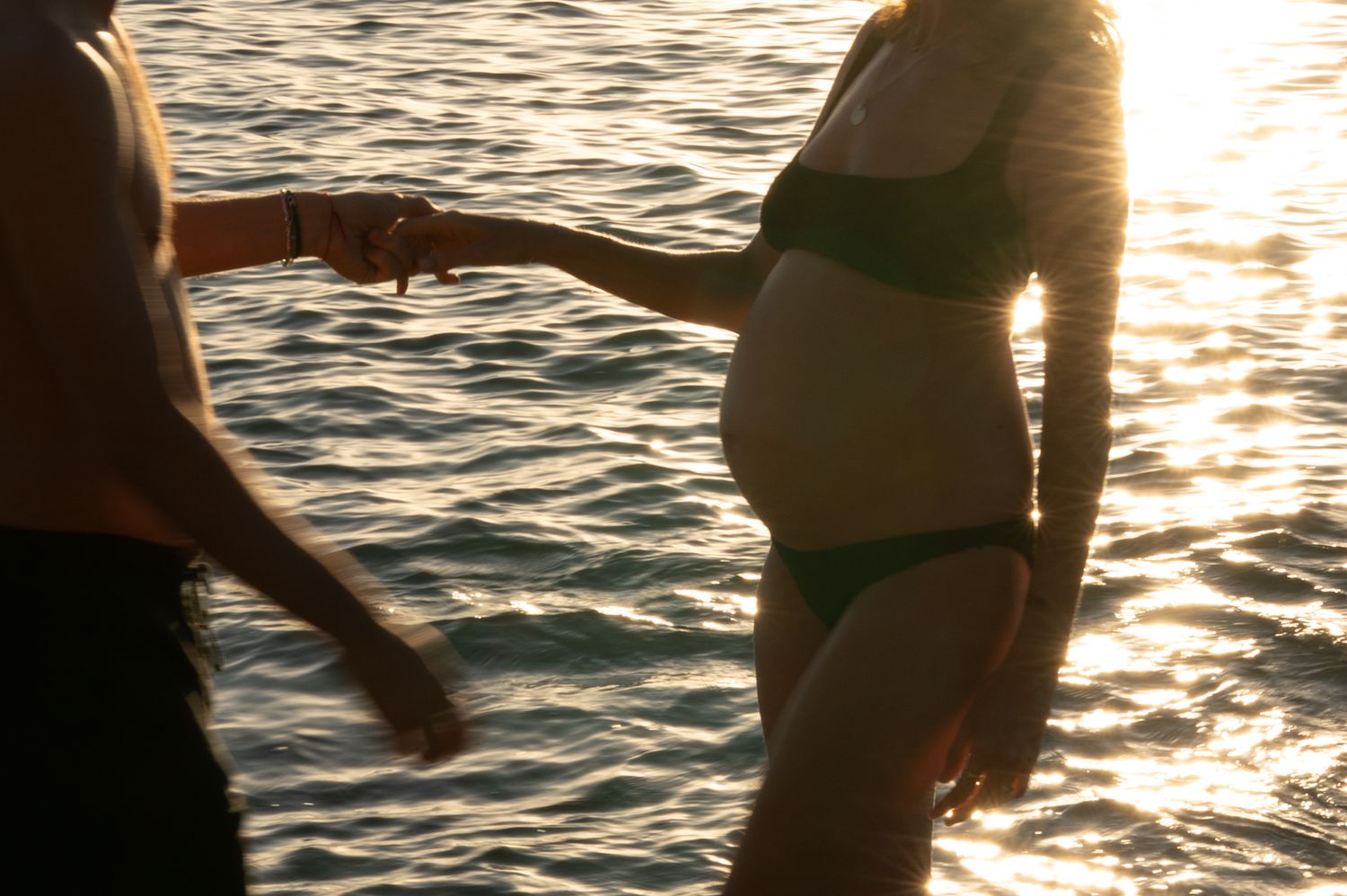 Pregnant woman in green bikini holding partner's hand while standing in sunlit shimmering ocean water. Maternity Photography
