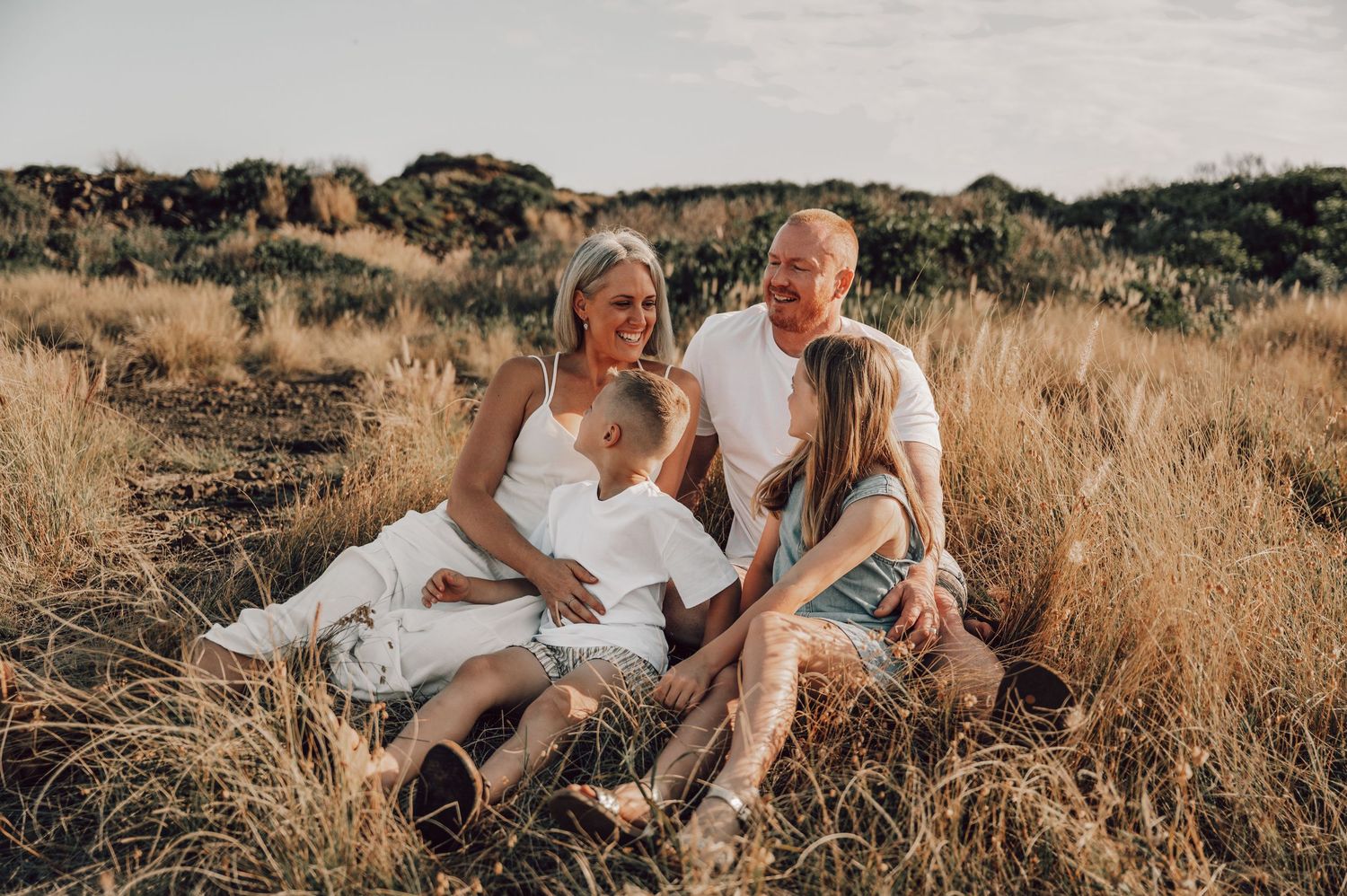 Happy family of four sitting together in golden dry grass field during warm sunset light photography session. Family Photography