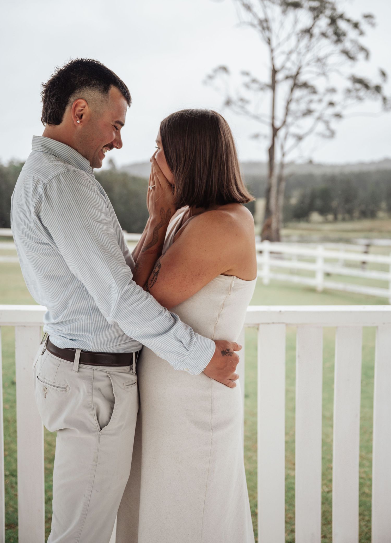 Couple embracing on a white fence porch, woman covering her mouth in surprise, outdoor engagement photo.
Proposal Photography