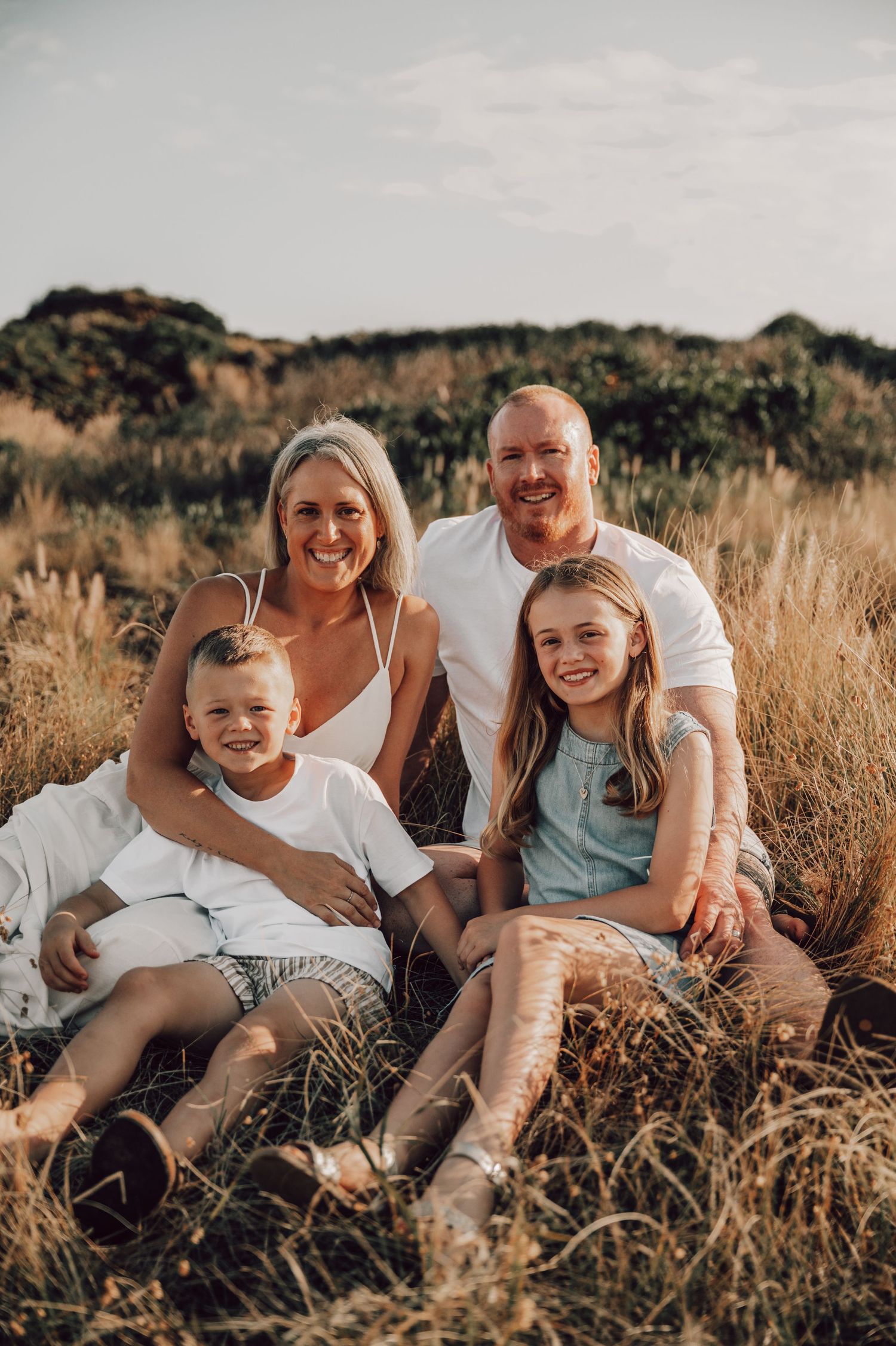Happy family of four sitting together in golden grass field, smiling outdoors during sunset in white casual outfits. Family Photography