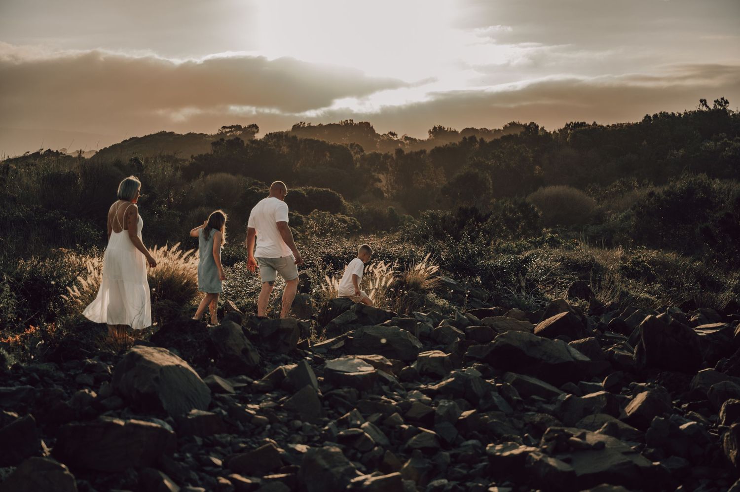 Family walking on rocky terrain at golden sunset with lush greenery and dramatic cloudy sky in background. Family Photography