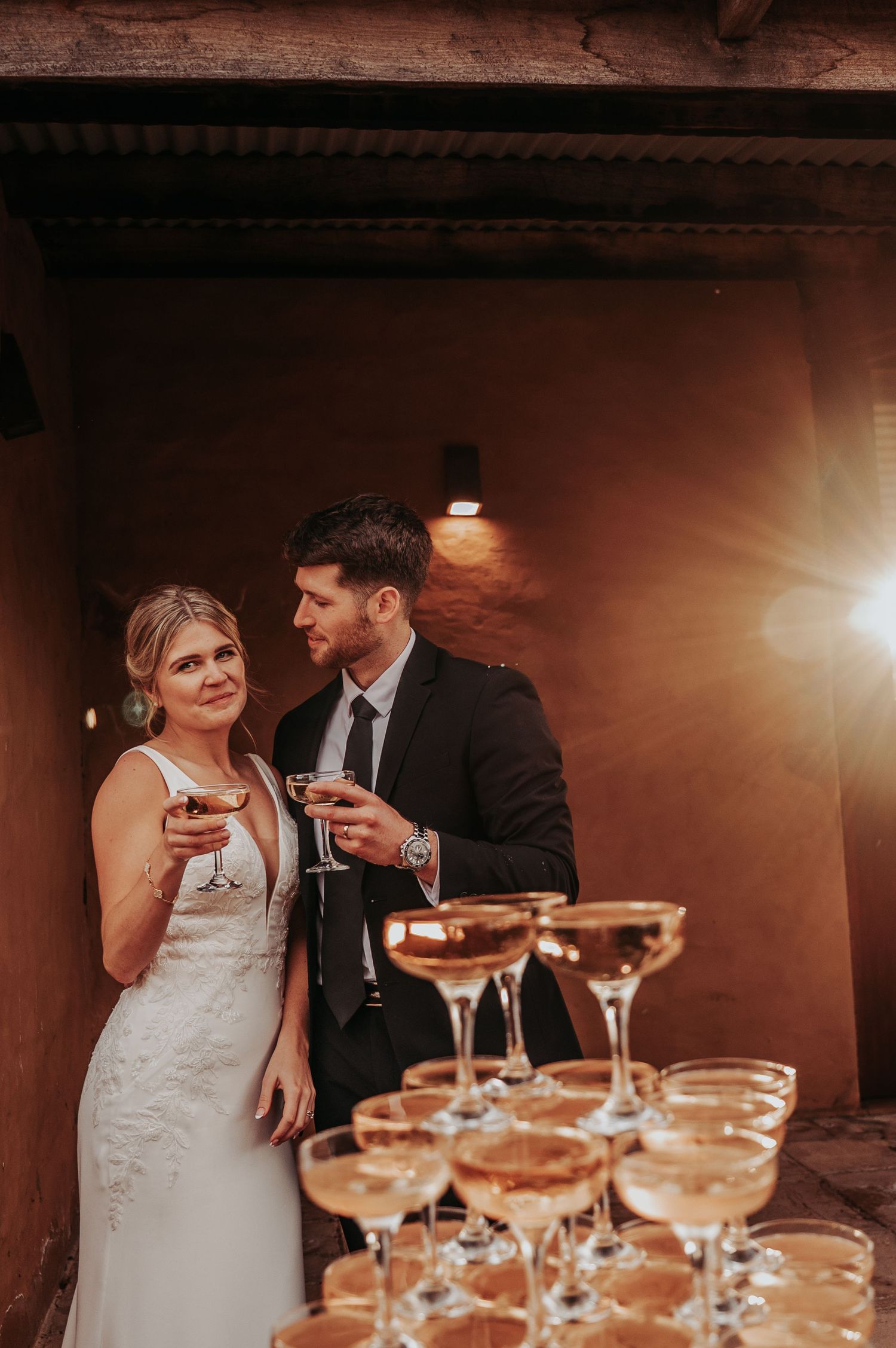 Bride and groom holding champagne coupe glasses beside a champagne tower at their wedding reception. Wedding Photography, Champagne tower