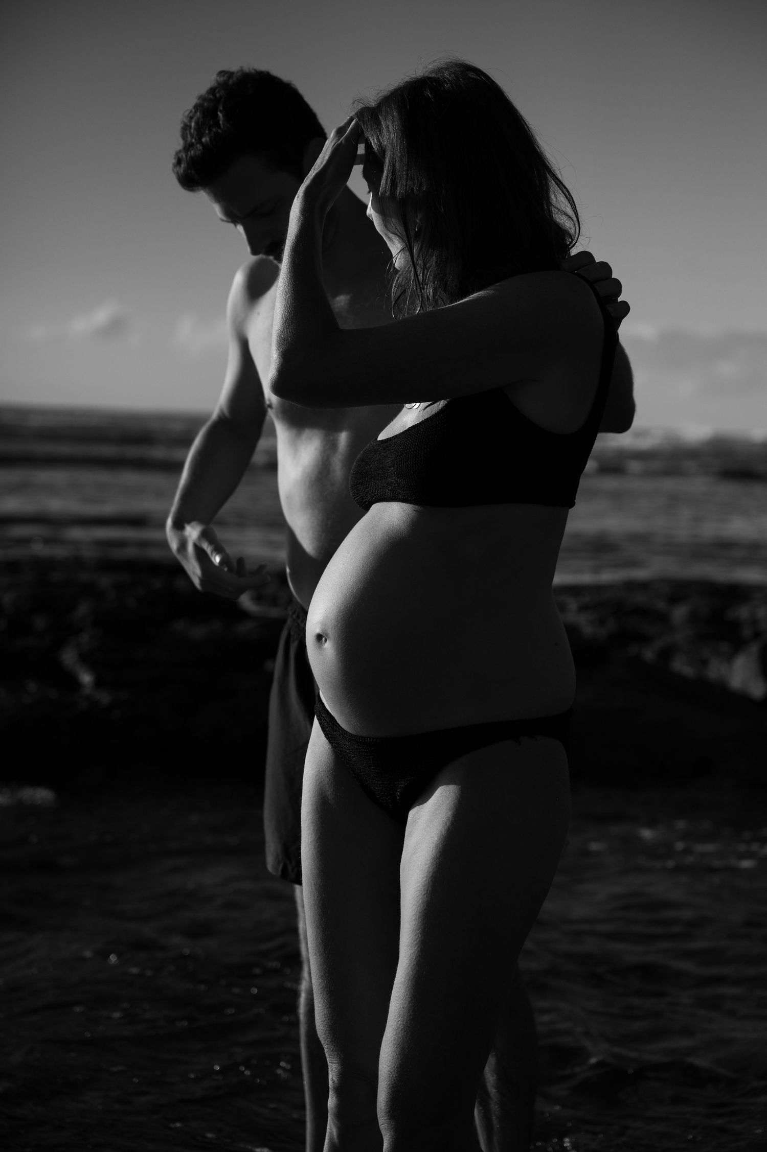 Pregnant couple embracing on rocky beach in black and white maternity photography session. Maternity Photography