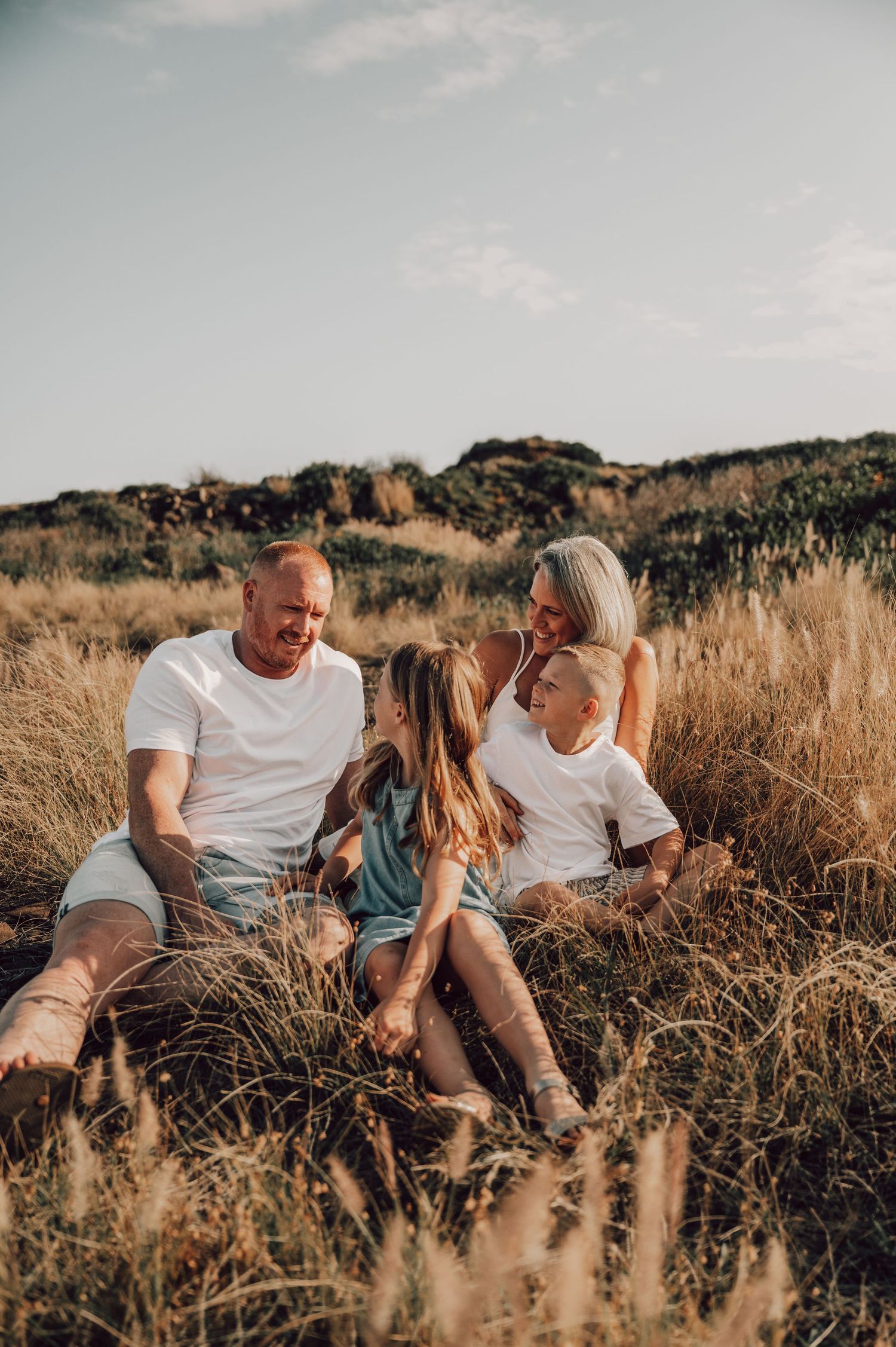 Family of four sitting together in golden grass field, parents and two children wearing white shirts, smiling outdoors. Family Photography