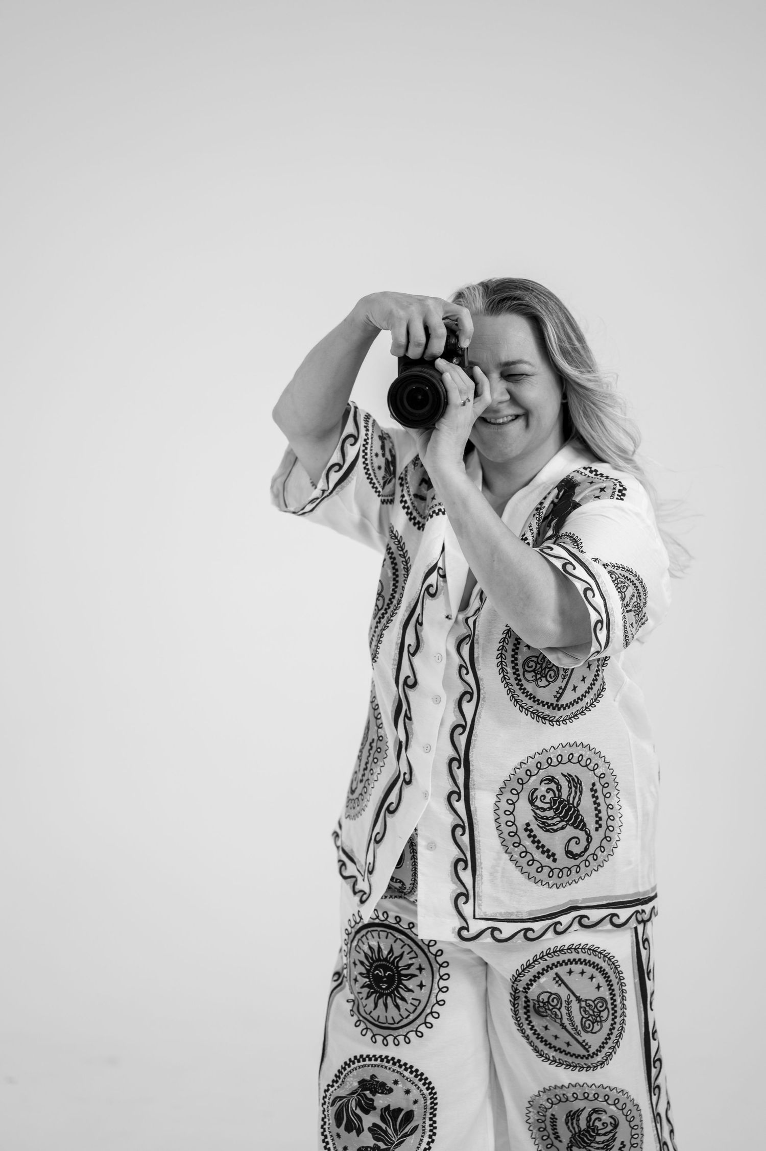 Black and white portrait of smiling female photographer holding DSLR camera in patterned white outfit. Photographer