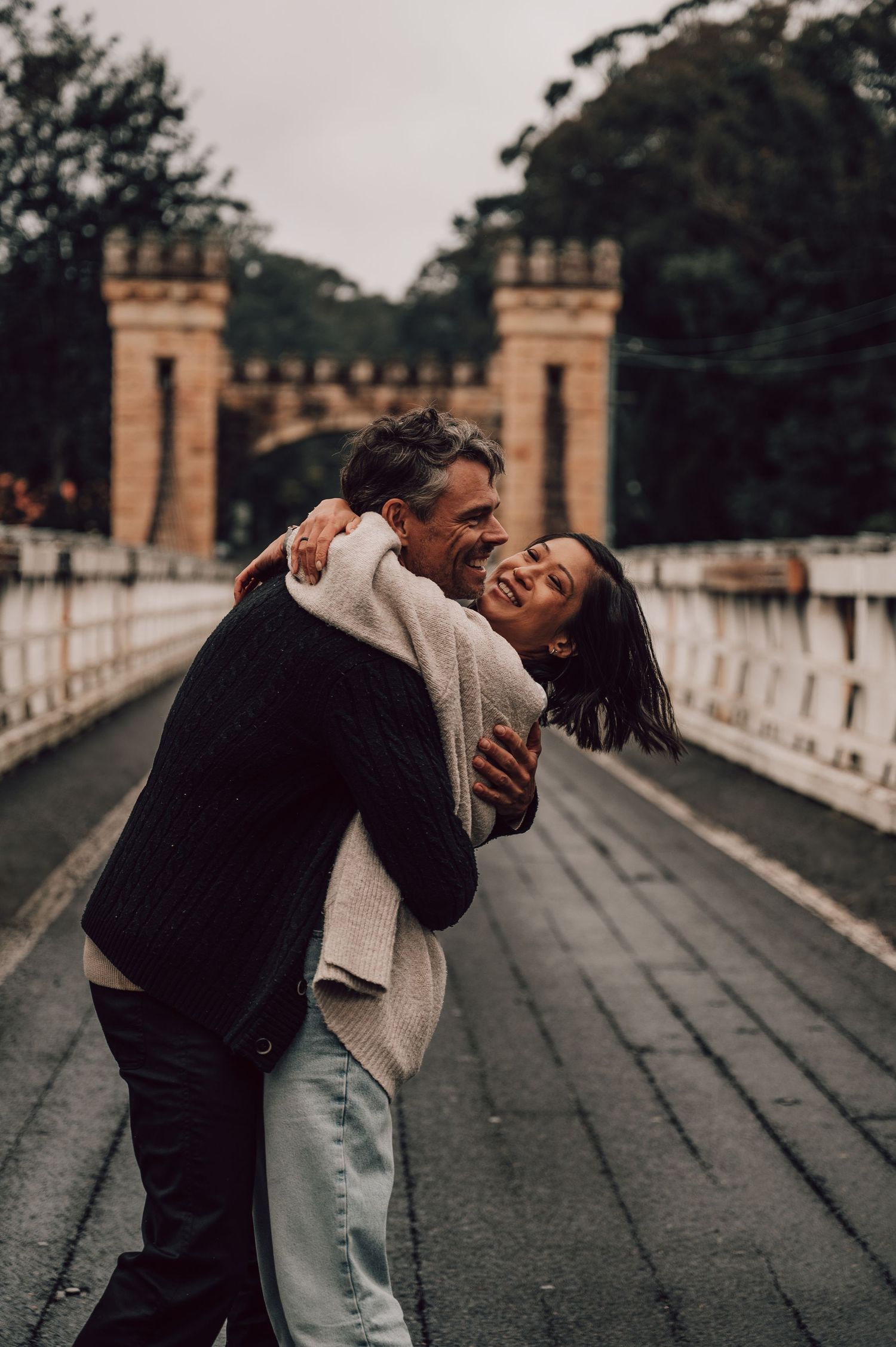 Happy couple embracing and laughing on a bridge with historic stone gatehouse towers in background. Couples Photography