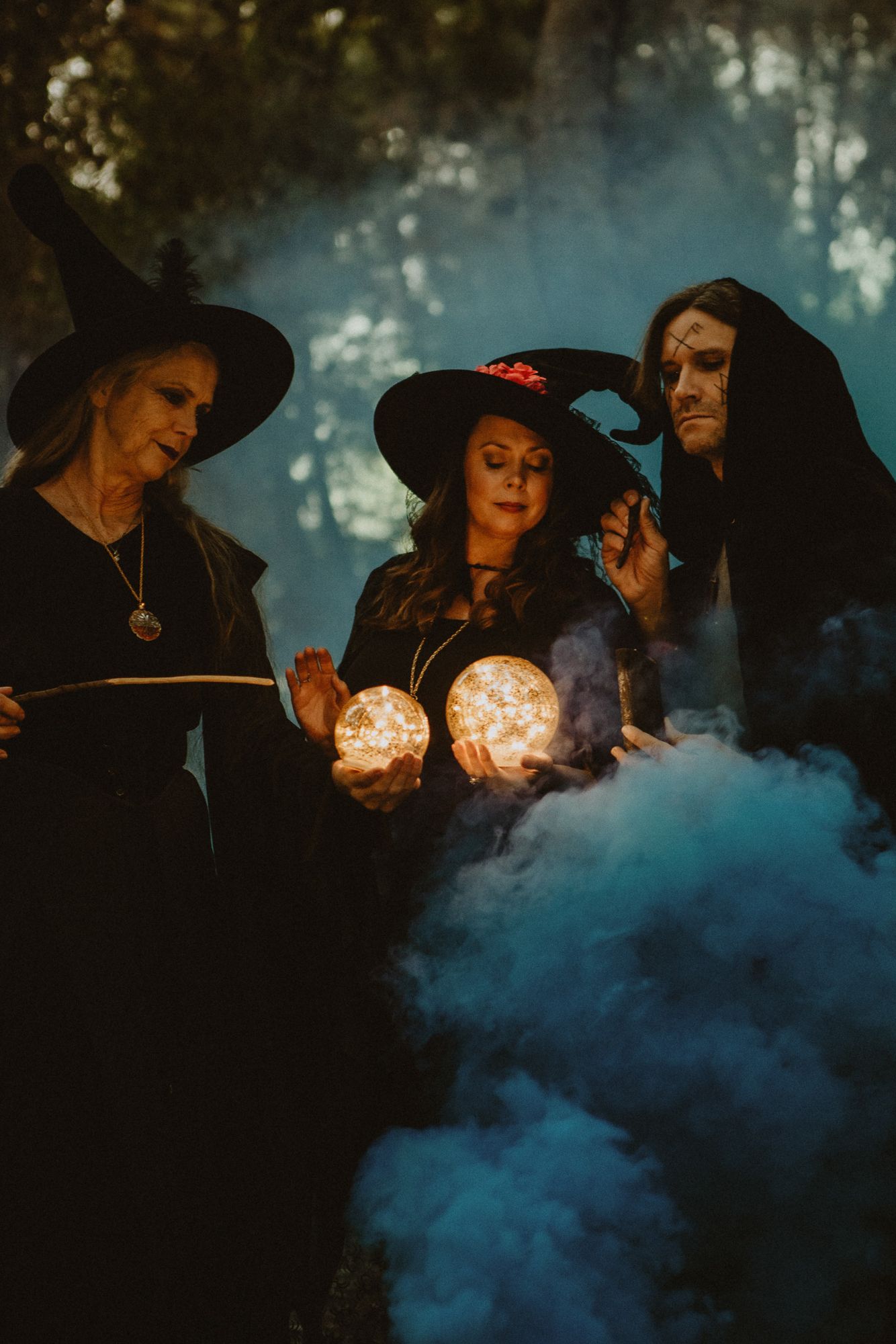 Three witches pose with glowing orbs and smoke at twilight
