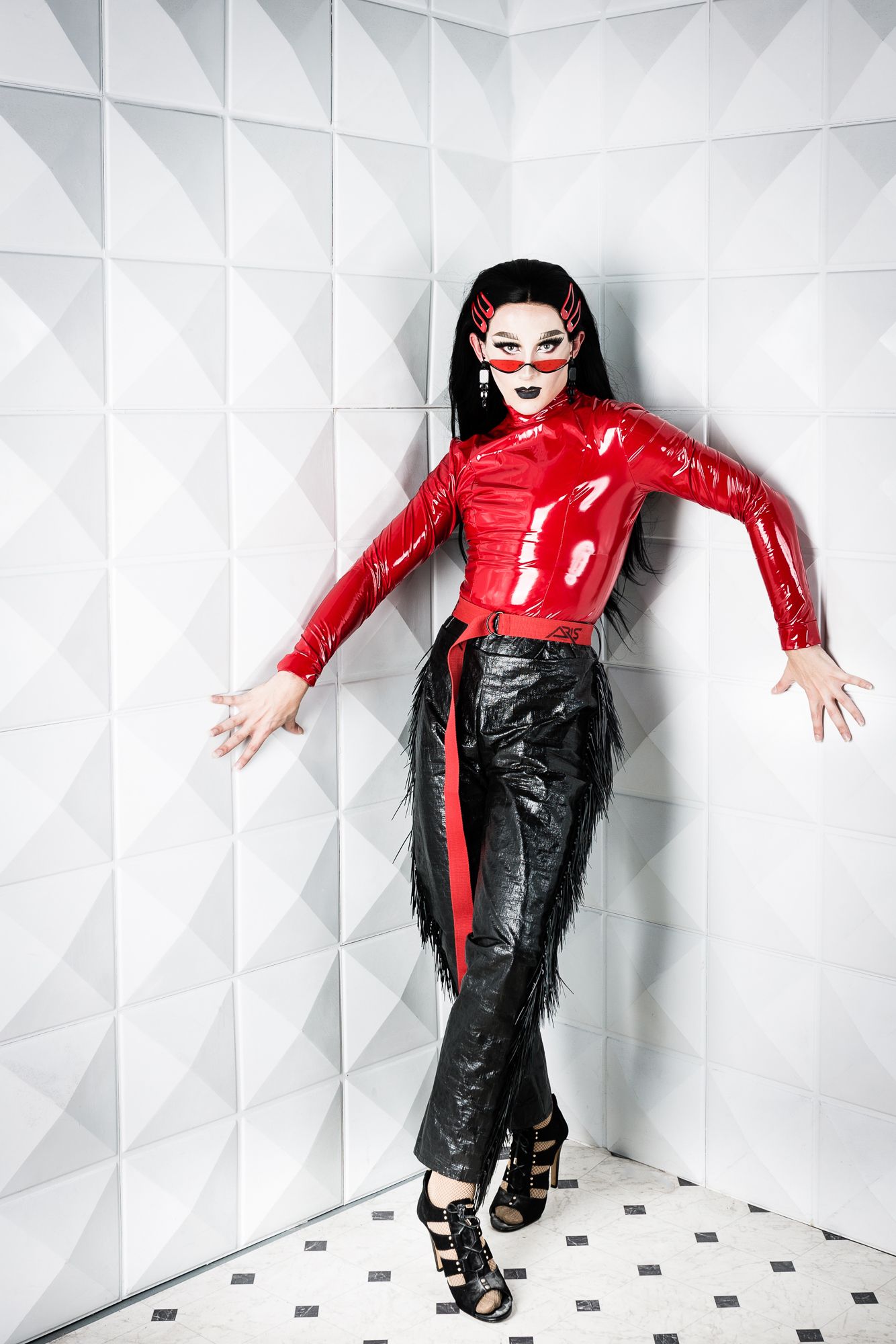 A drag queen in self-made costume poses in the corner of a white set