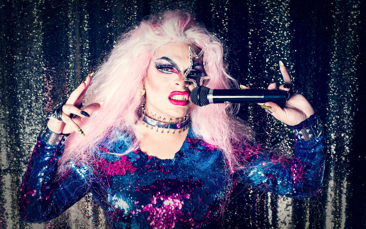 A drag queen dressed as an '80s performer poses with a microphone in front of a glittery background