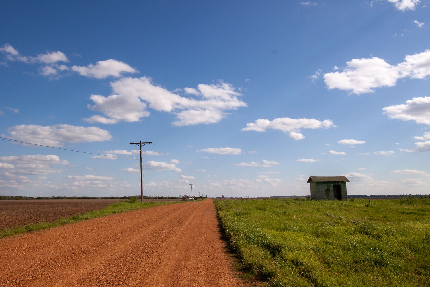 A red dirt road runs alongside a field under a blue sky with puffy clouds in Mississippi