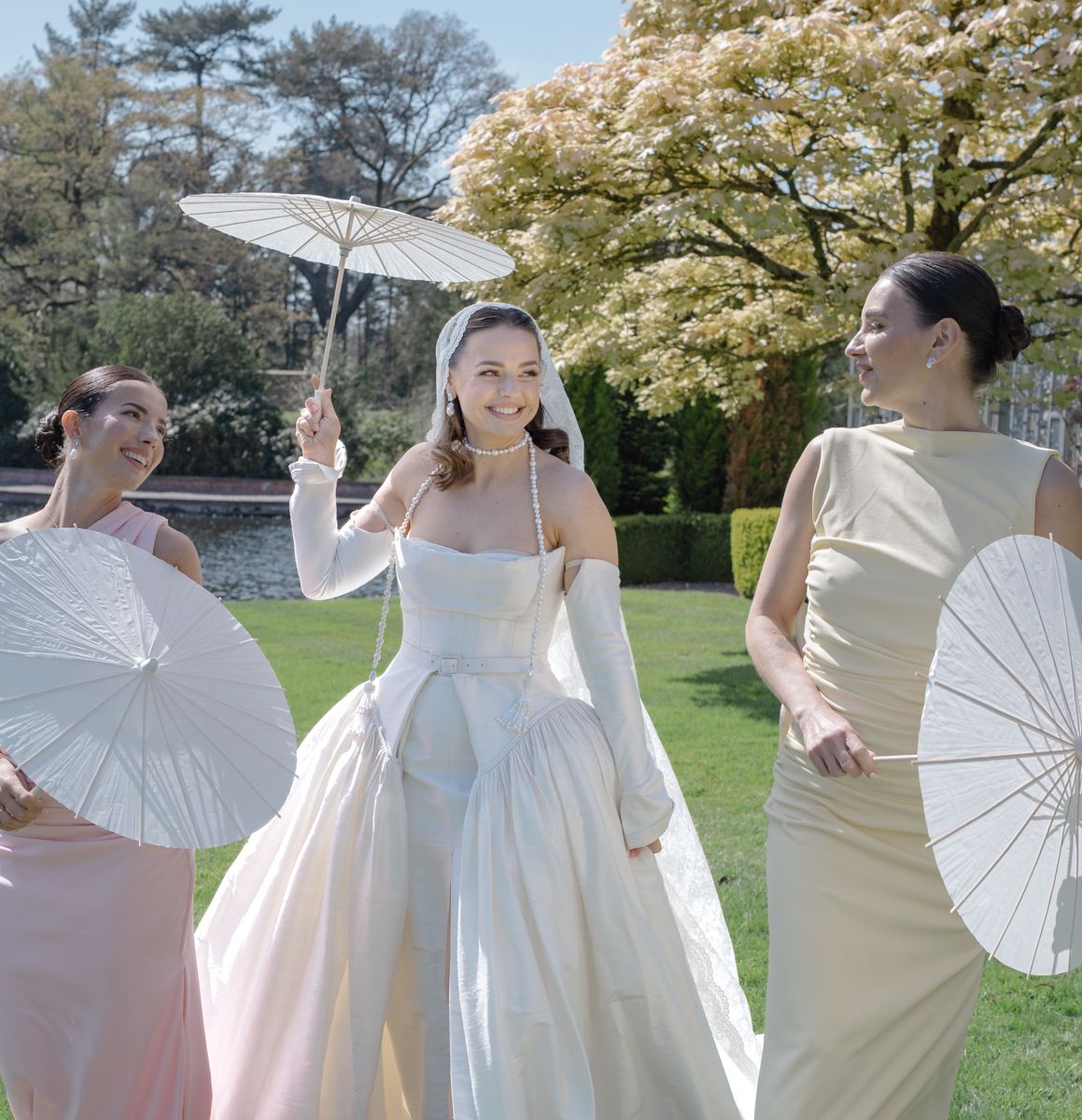 Bride and bridesmaids walking the grounds of Capesthorne Hall with sun parasol and long veil - editorial wedding photographer Cheshire