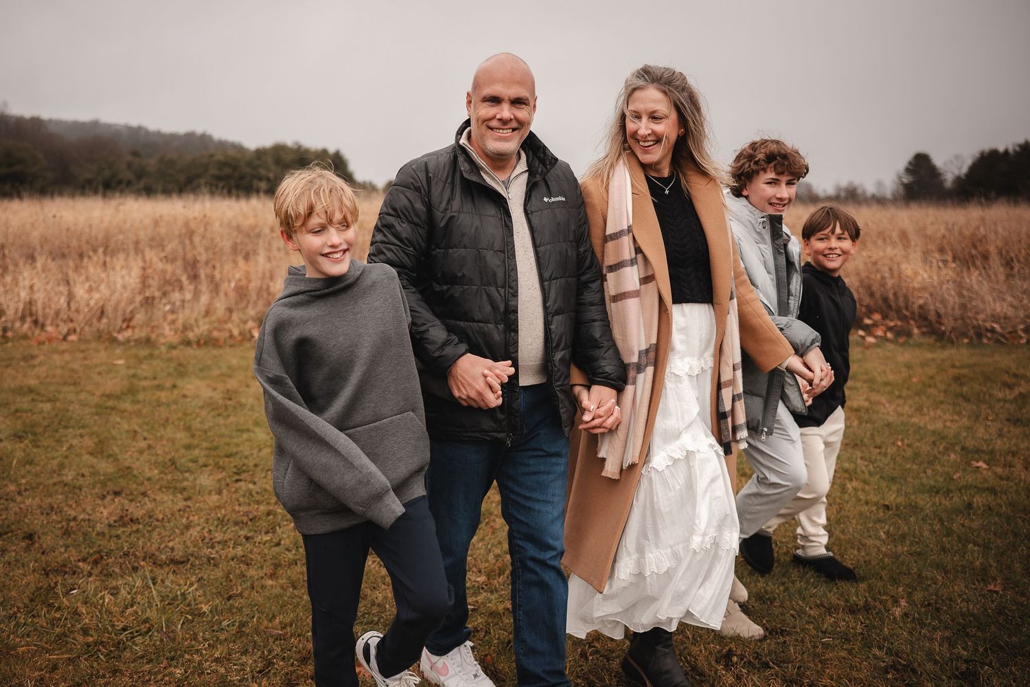 A group of people stand together outdoors in a field during autumn with tall grass and trees in the background.