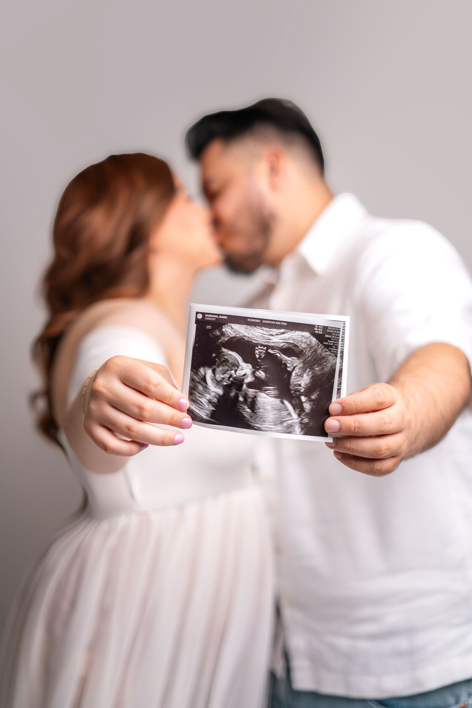 Couple holding ultrasound photo while sharing intimate moment against grey background.