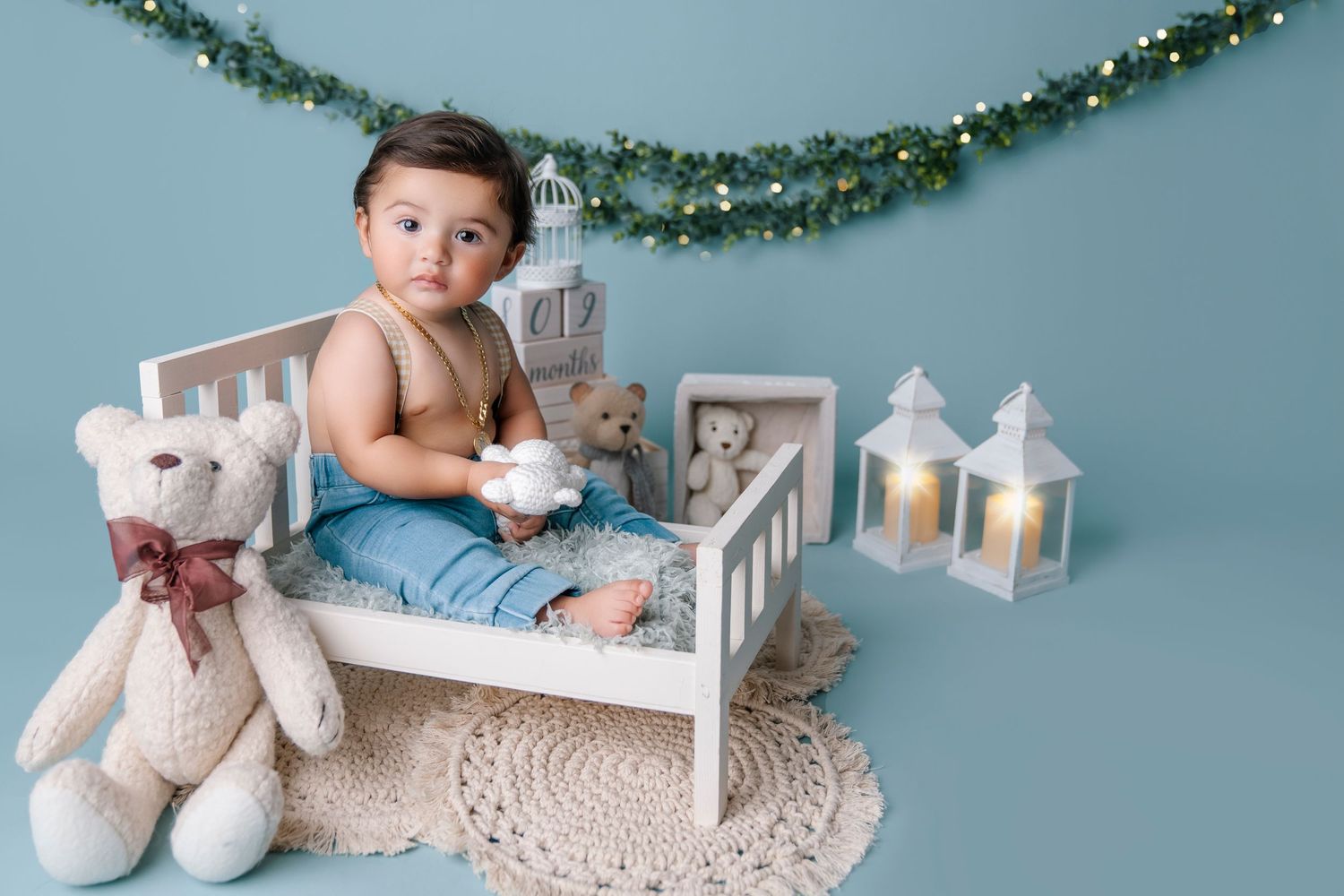 Baby sits on small white bed with teddy bear and lanterns in holiday setting.
