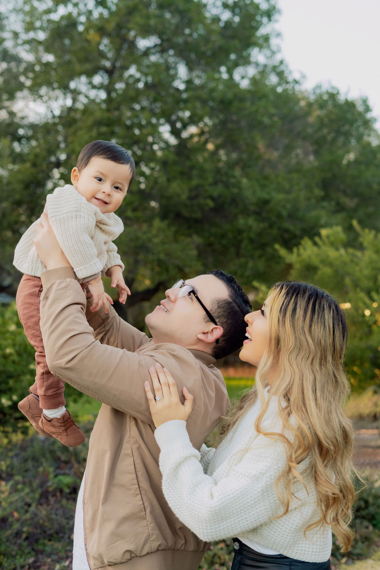 Family shares joyful moment outdoors lifting baby in white sweater against green background.