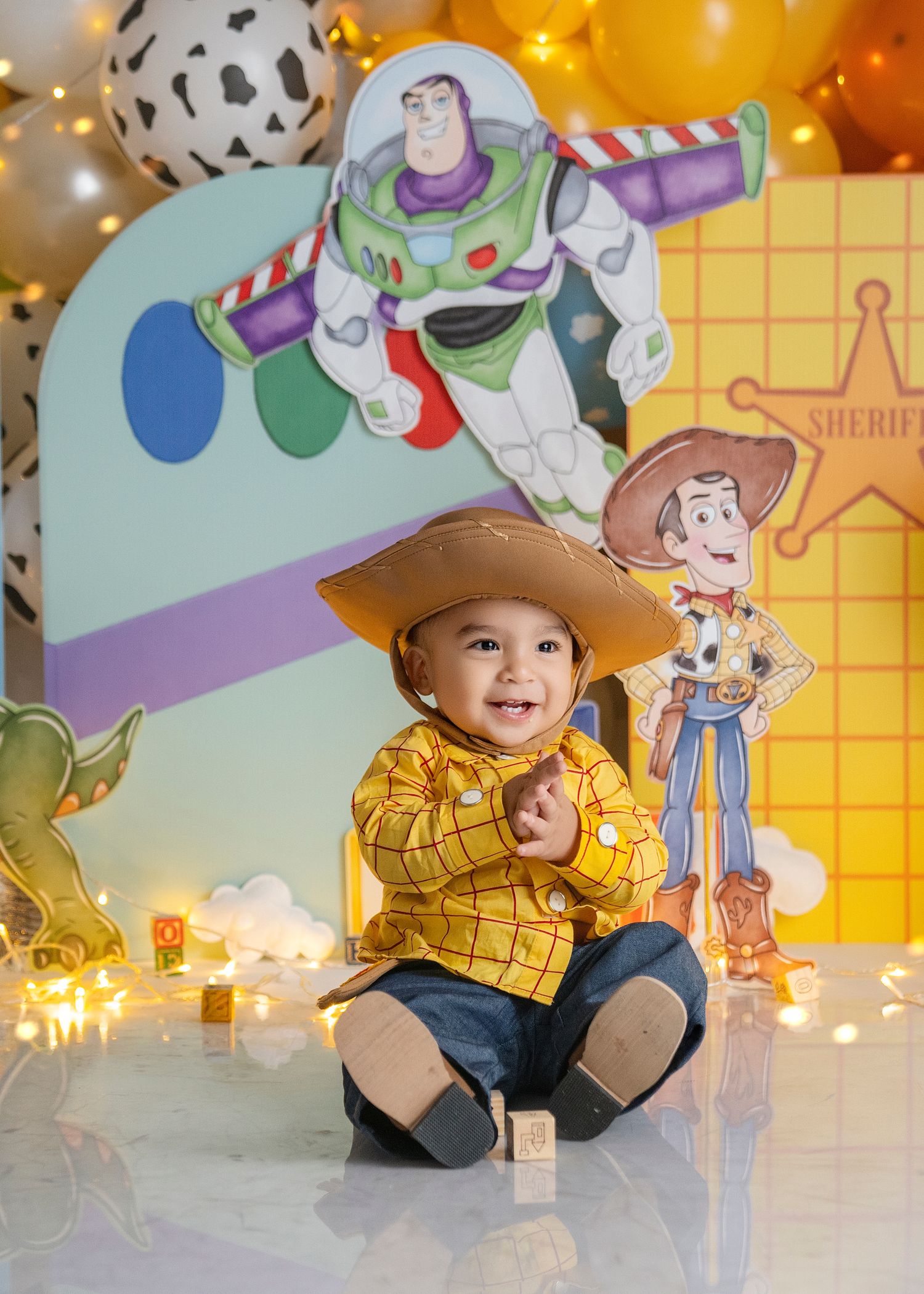 Smiling baby dressed as Woody from Toy Story sits before a colorful Toy Story themed backdrop with balloons.