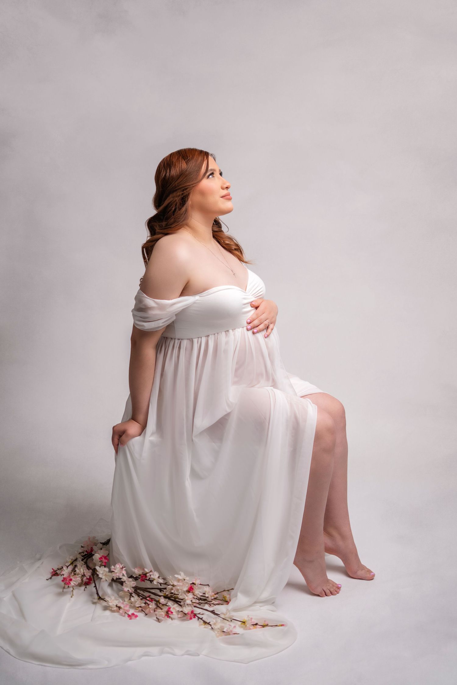 Ethereal maternity portrait of woman in flowing white dress against light backdrop.