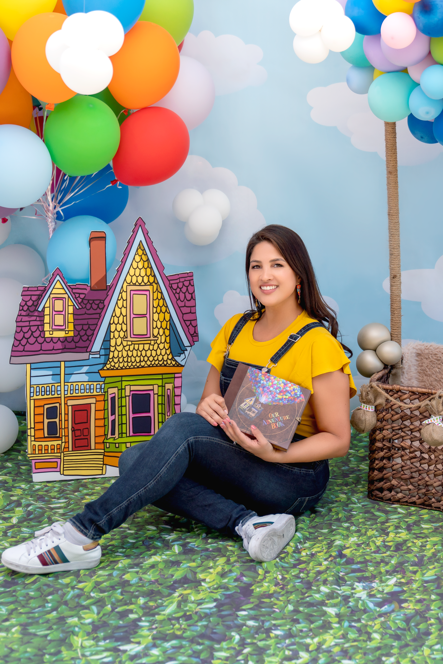 Whimsical photo setup with colorful balloons and illustrated house backdrop against blue sky background.
