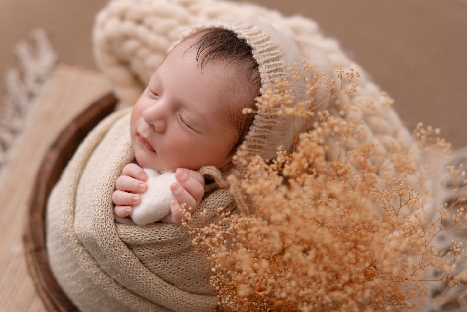 Sleeping baby wrapped in knit blanket with dried flowers and beige backdrop.