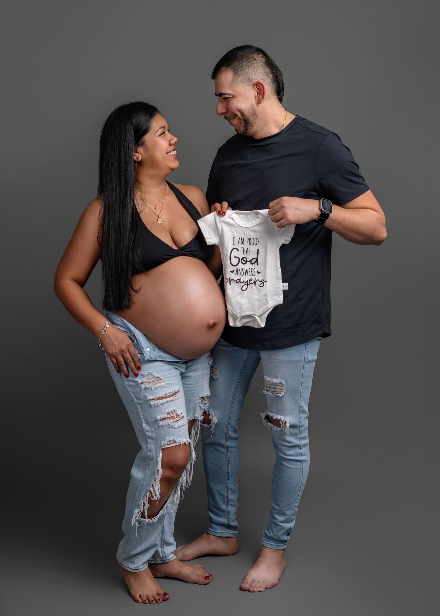 Expectant couple holding baby onesie during maternity photoshoot against grey background.