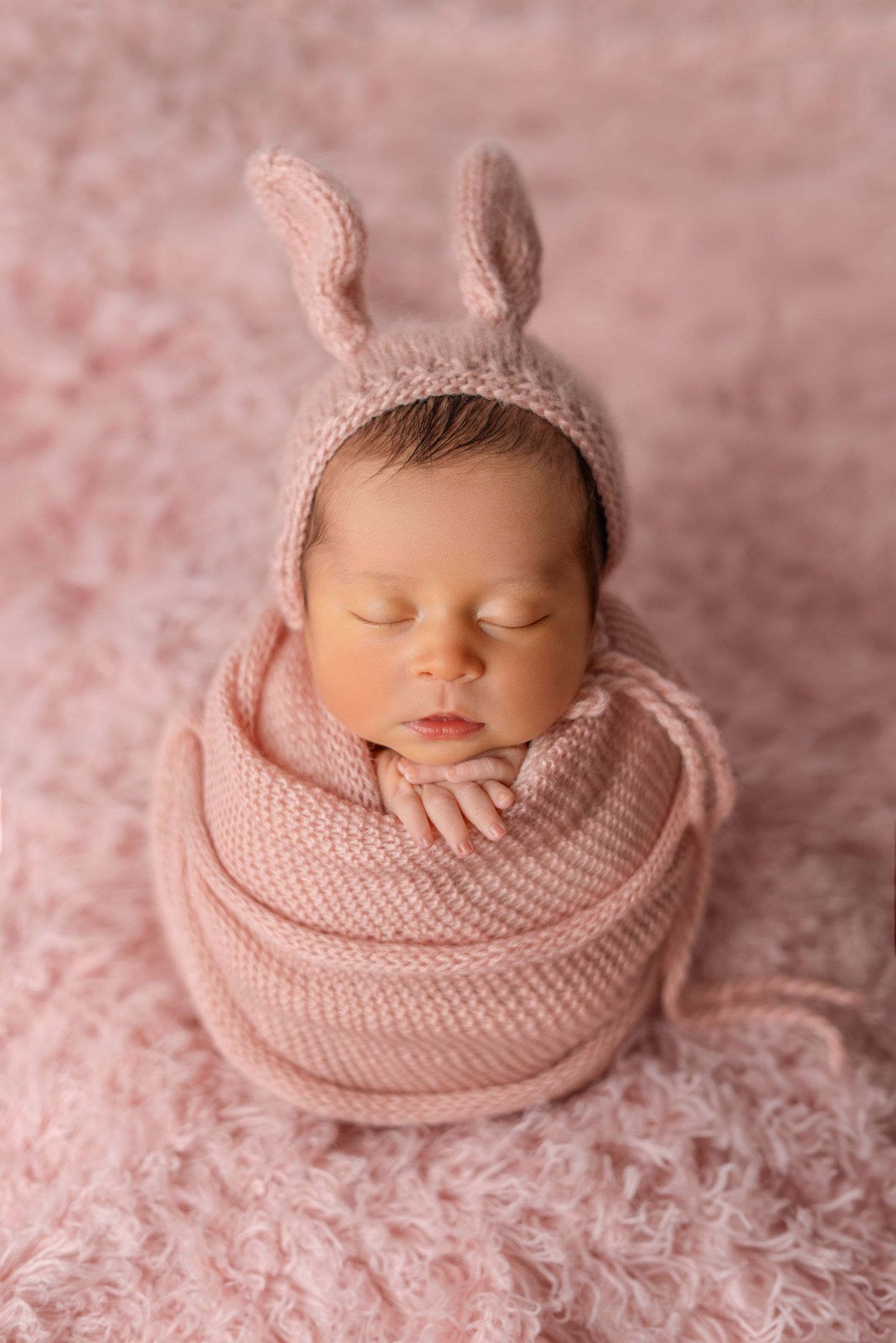 Sleeping newborn wrapped in pink knit blanket wearing bunny ear bonnet on textured background.