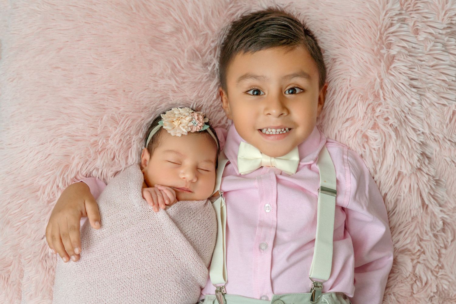 Siblings pose together on pink furry backdrop with newborn in floral headband.
