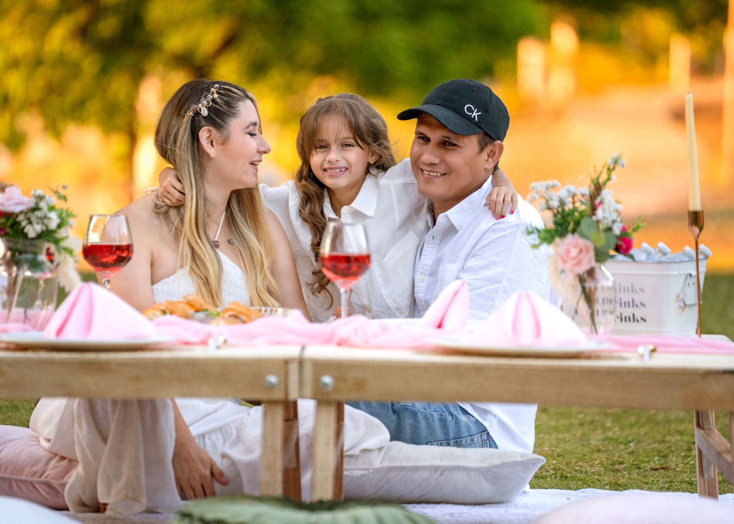 A family enjoys a romantic outdoor picnic with wine and flowers at sunset.