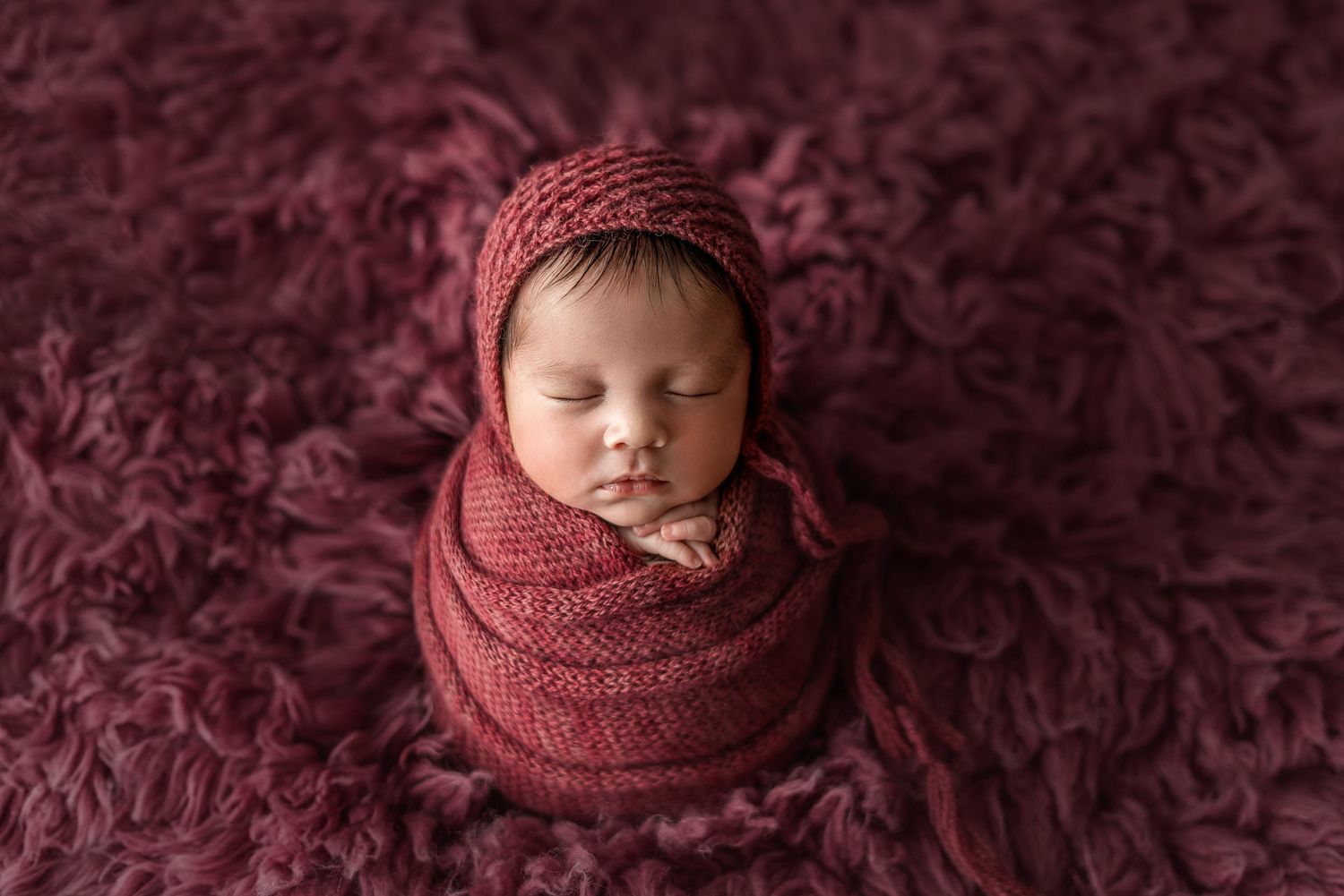 Newborn wrapped in burgundy knit swaddle on matching textured background.