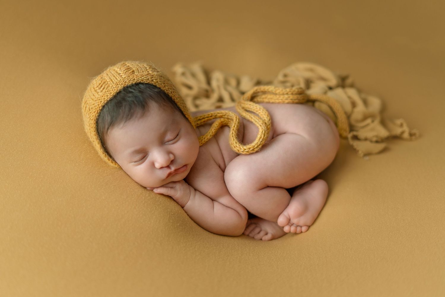 Sleeping newborn wearing yellow bonnet on golden backdrop.