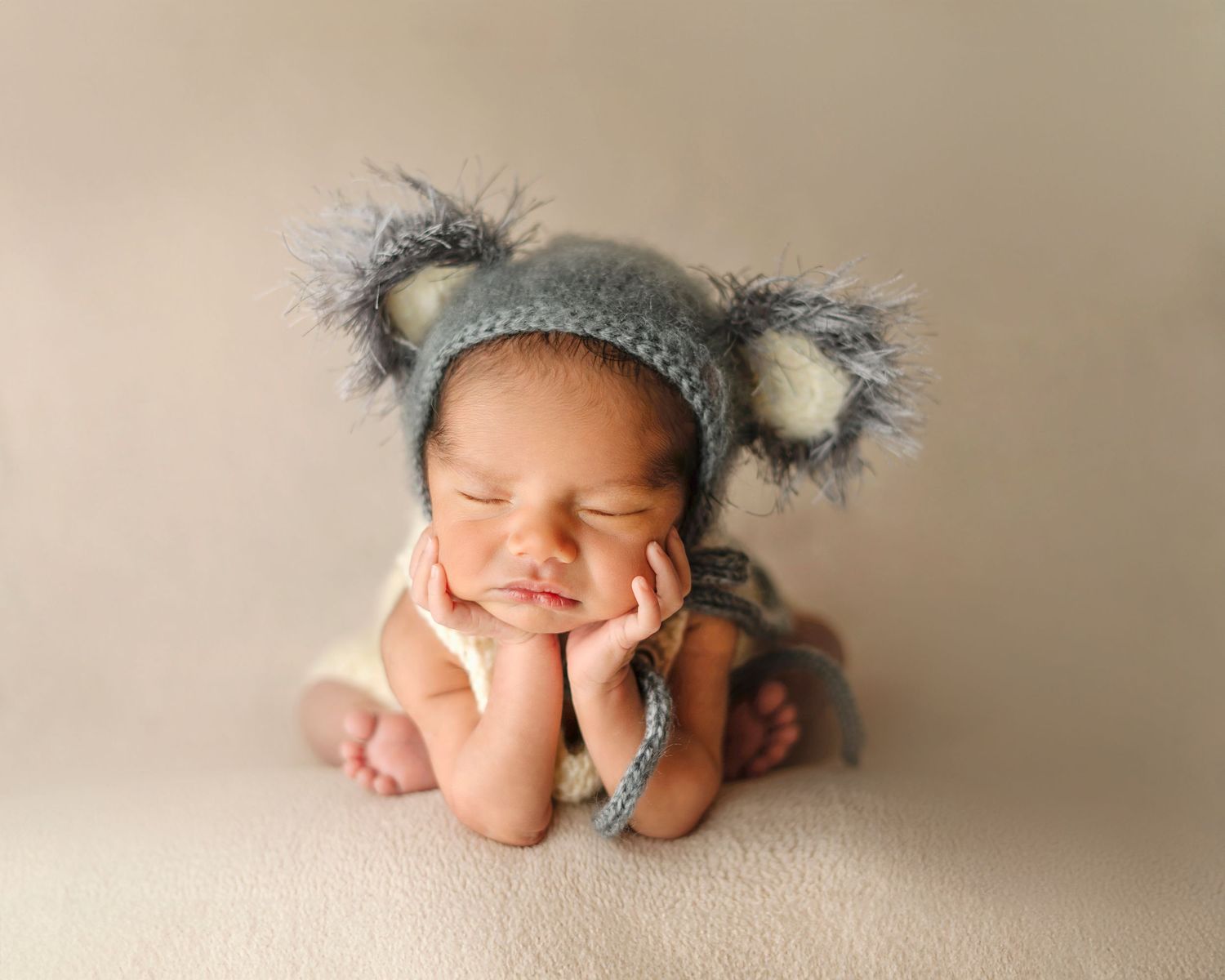 Sleeping baby wearing gray koala hat poses with hands under chin on beige background.