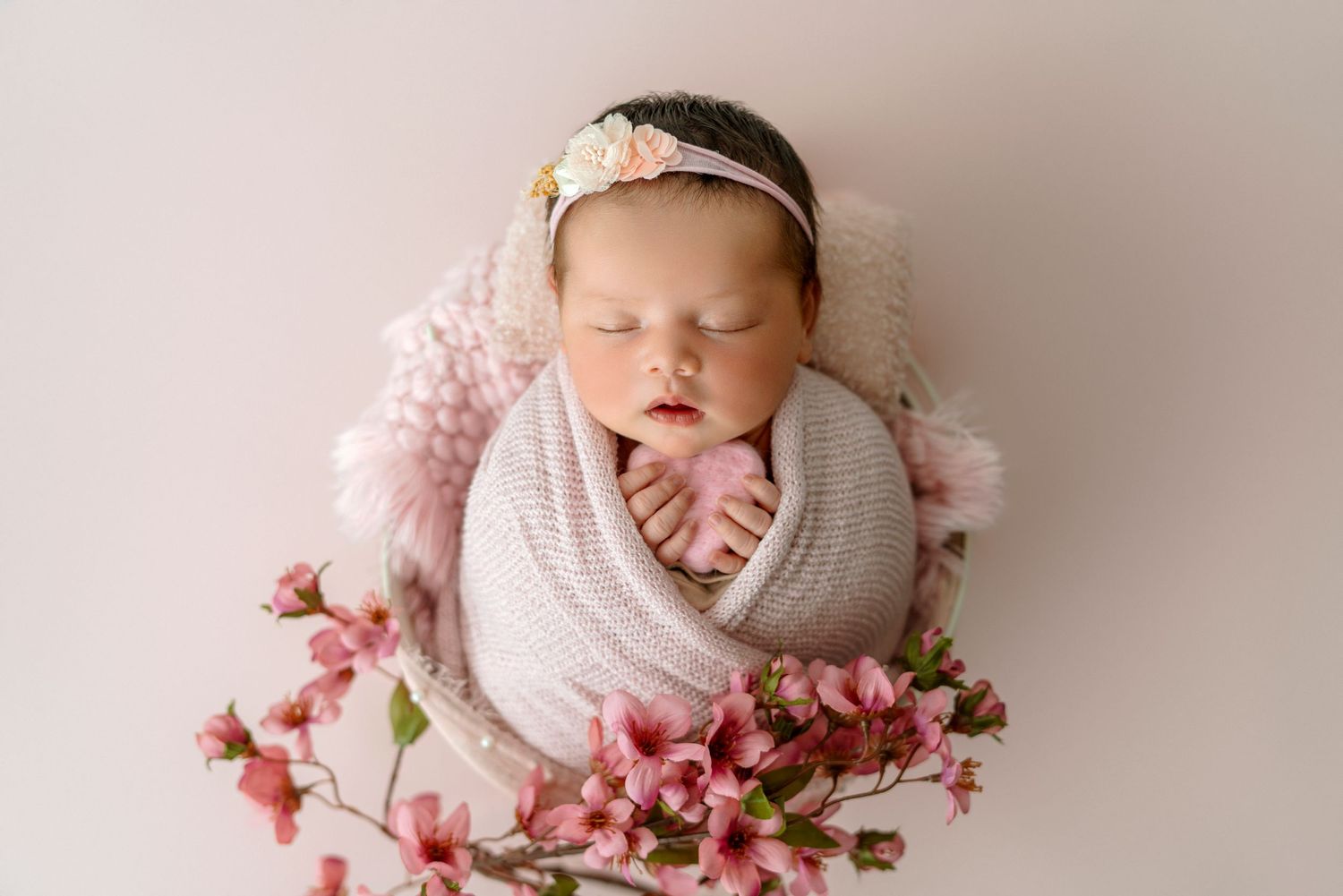Sleeping newborn wrapped in grey swaddle with pink flowers and headband on pink backdrop.