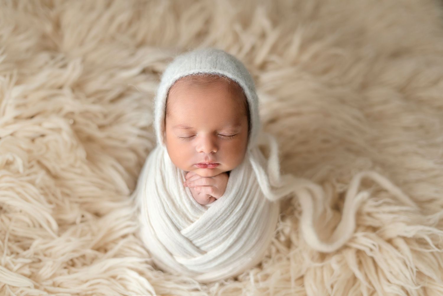 Peaceful newborn wrapped in white swaddle on textured cream background.