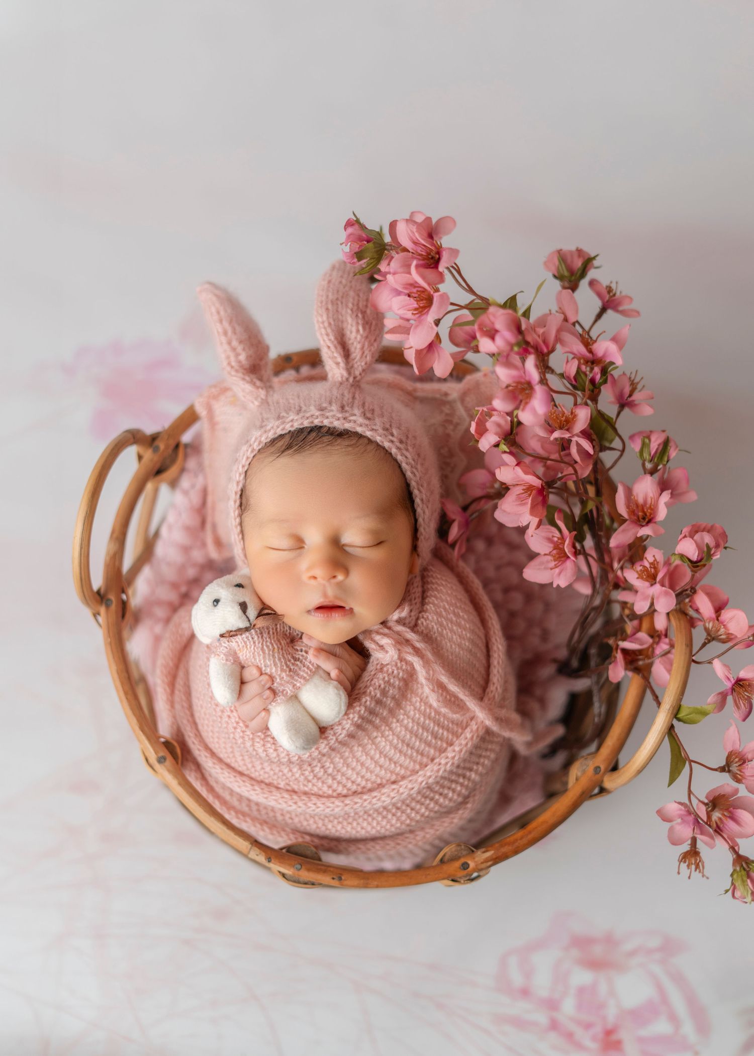 Newborn photography with pink flowers and bunny hat in rustic basket setting.