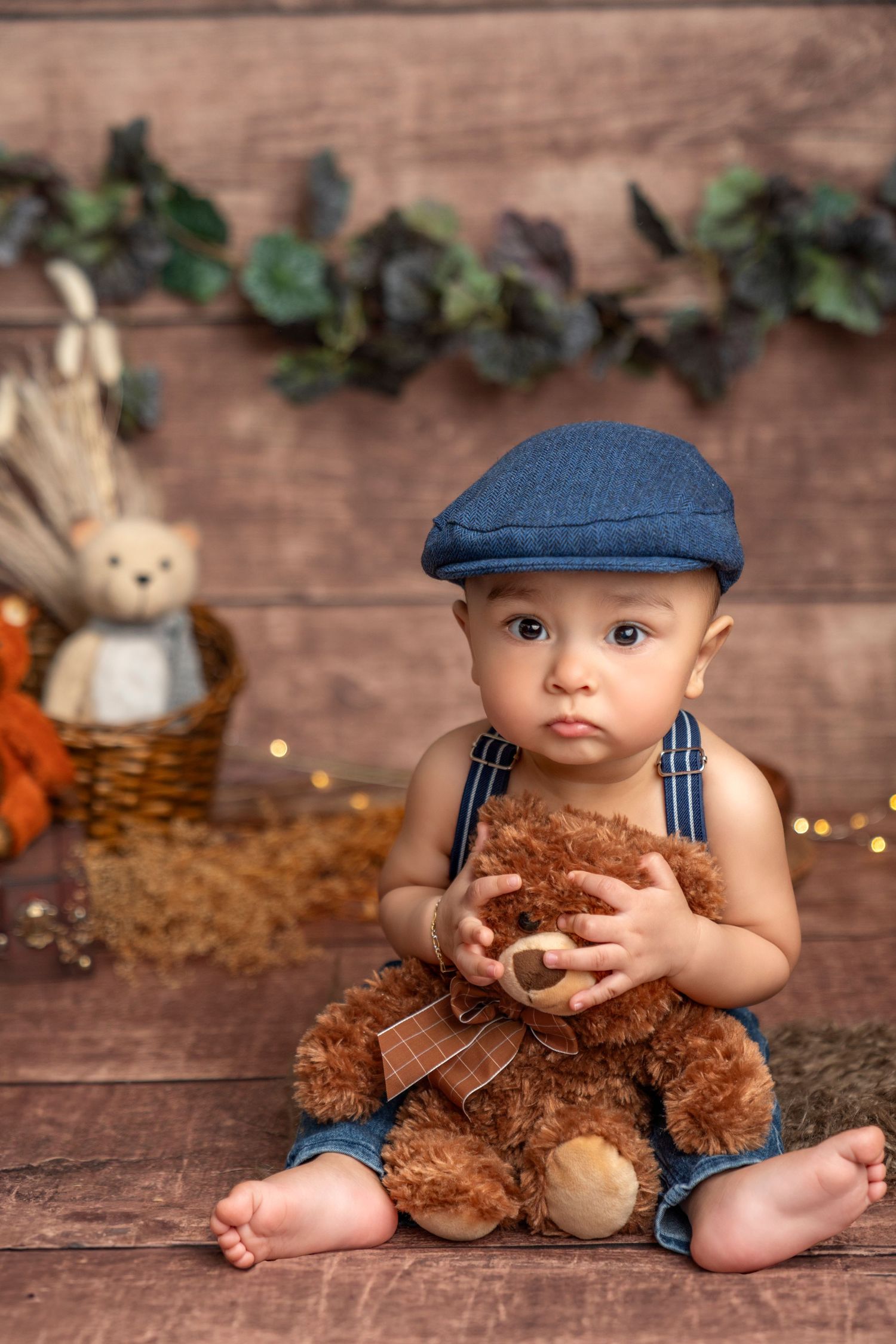 Baby in denim overalls and cap holds teddy bear against rustic wooden backdrop.