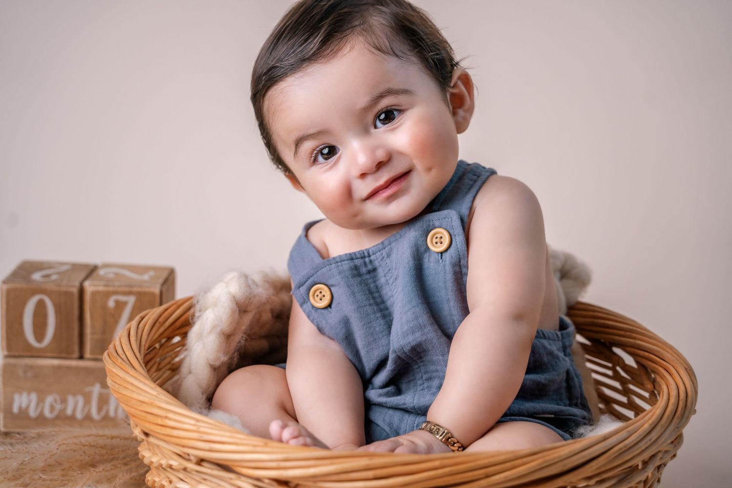 Baby in denim romper sitting in wicker basket with wooden blocks nearby.