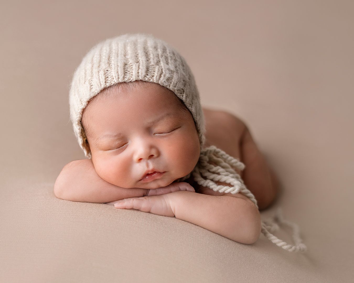 Sleeping newborn wearing knit bonnet on beige backdrop in studio setting.