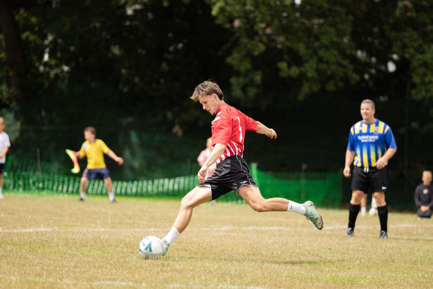 Soccer players in action during a match on a grass field with trees in background.
