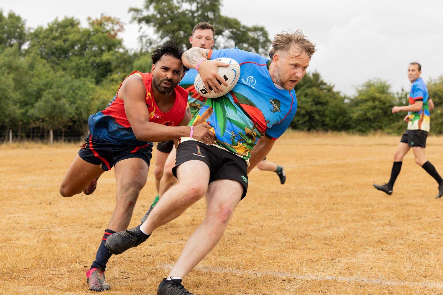 Rugby players battle for possession during an intense tackle on a dry field with trees in the background.
