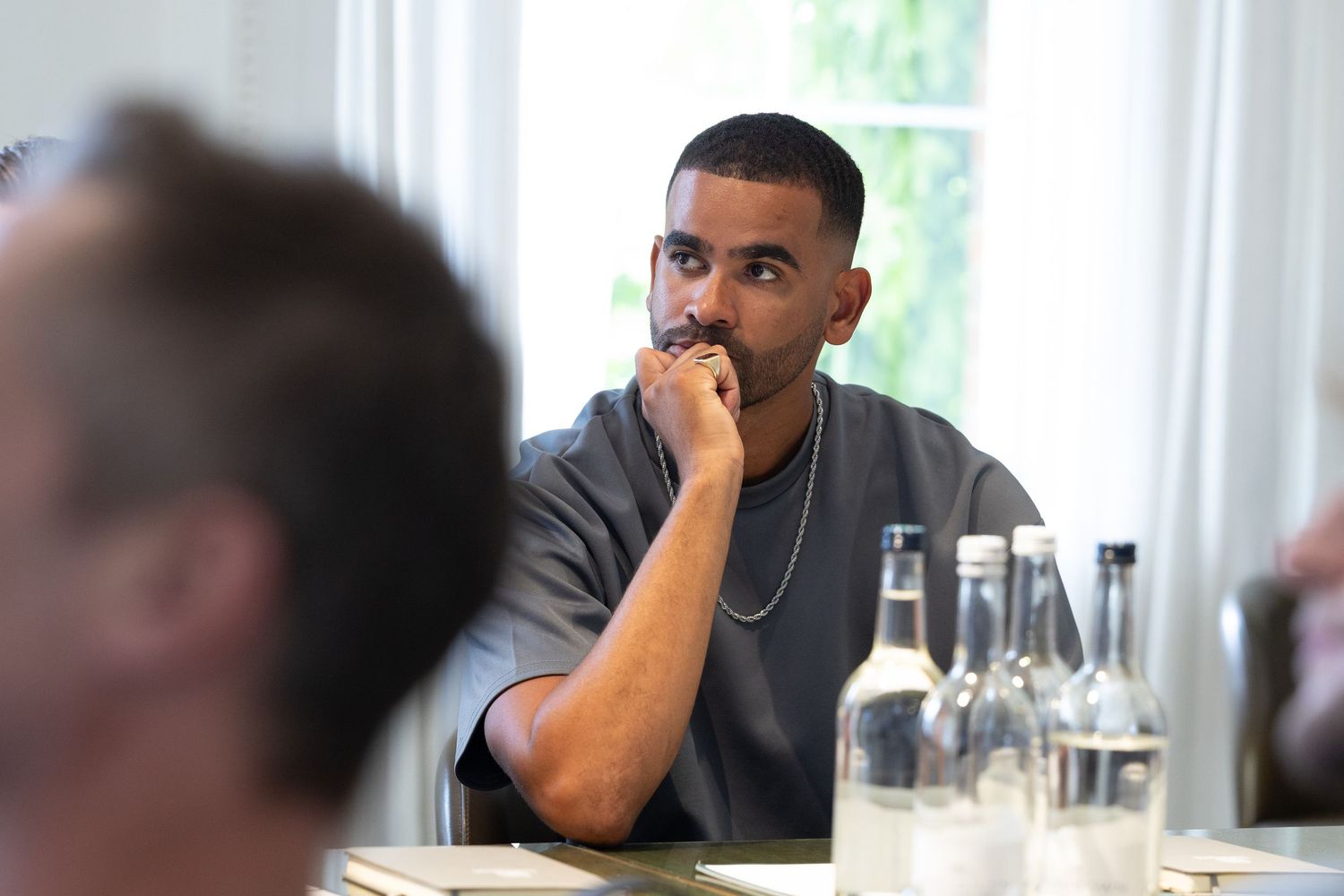 Person in grey shirt listening thoughtfully during meeting with bottles and glassware on table.