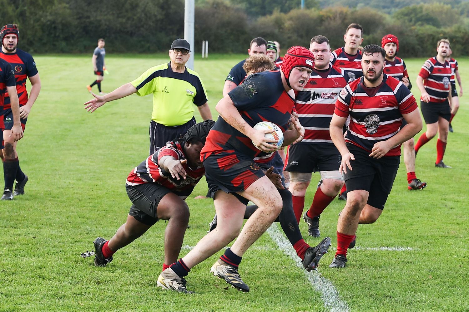 Rugby players engage in a tackle during a match on a grass field with players in red and black striped jerseys.