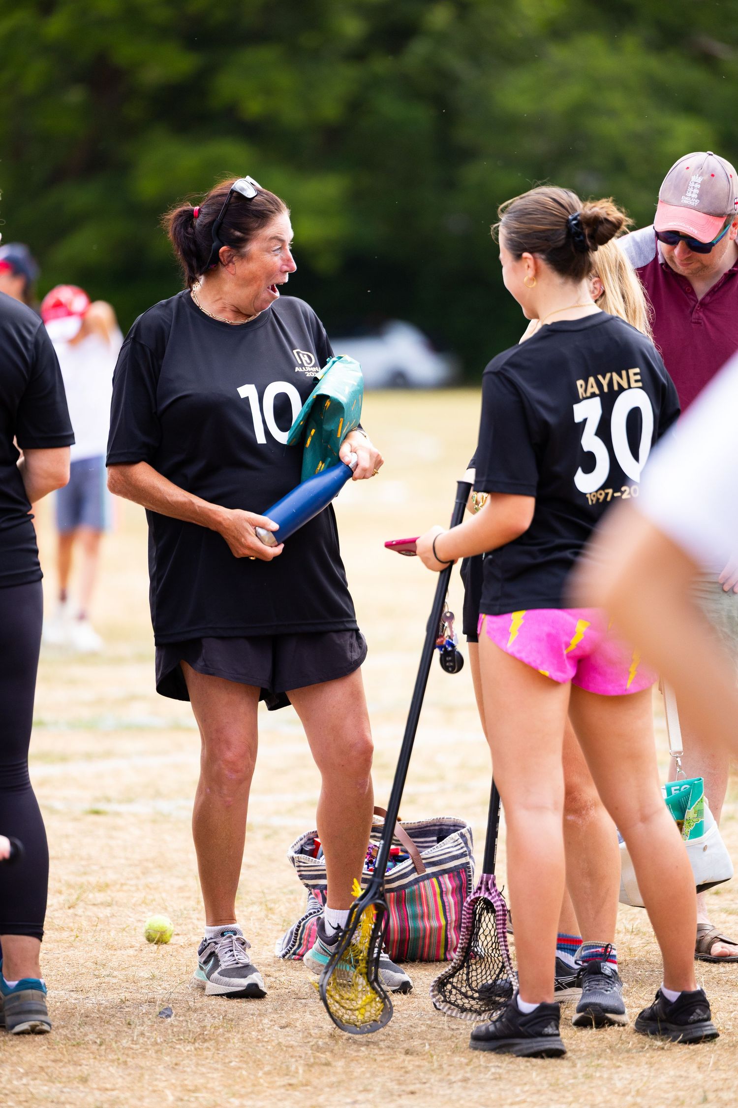 Female athletes in black jerseys chat while holding sports equipment on a dirt field during what appears to be a softball game.