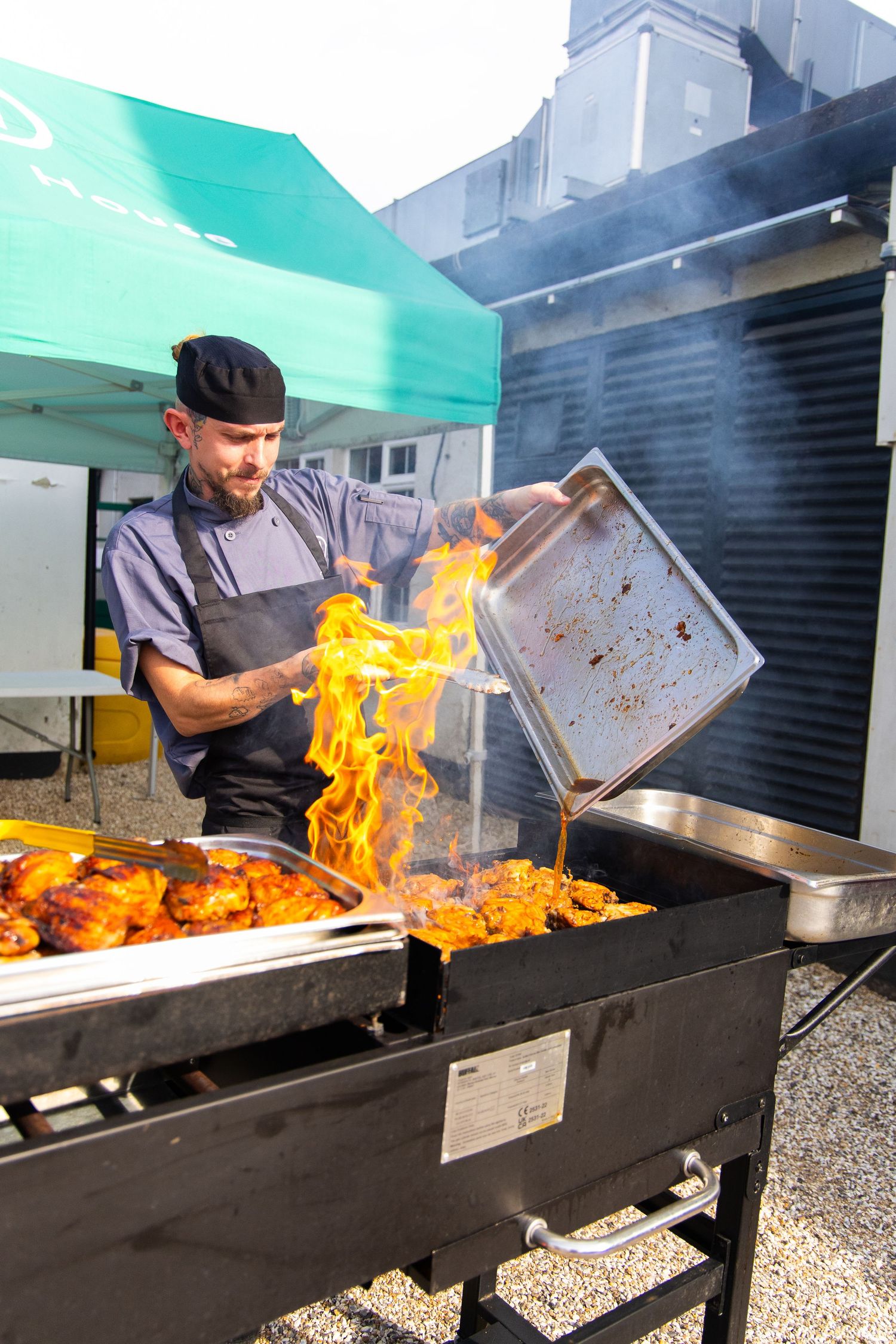 Chef grilling meat on outdoor barbecue grill with dramatic flames and smoke rising from cooking surface.