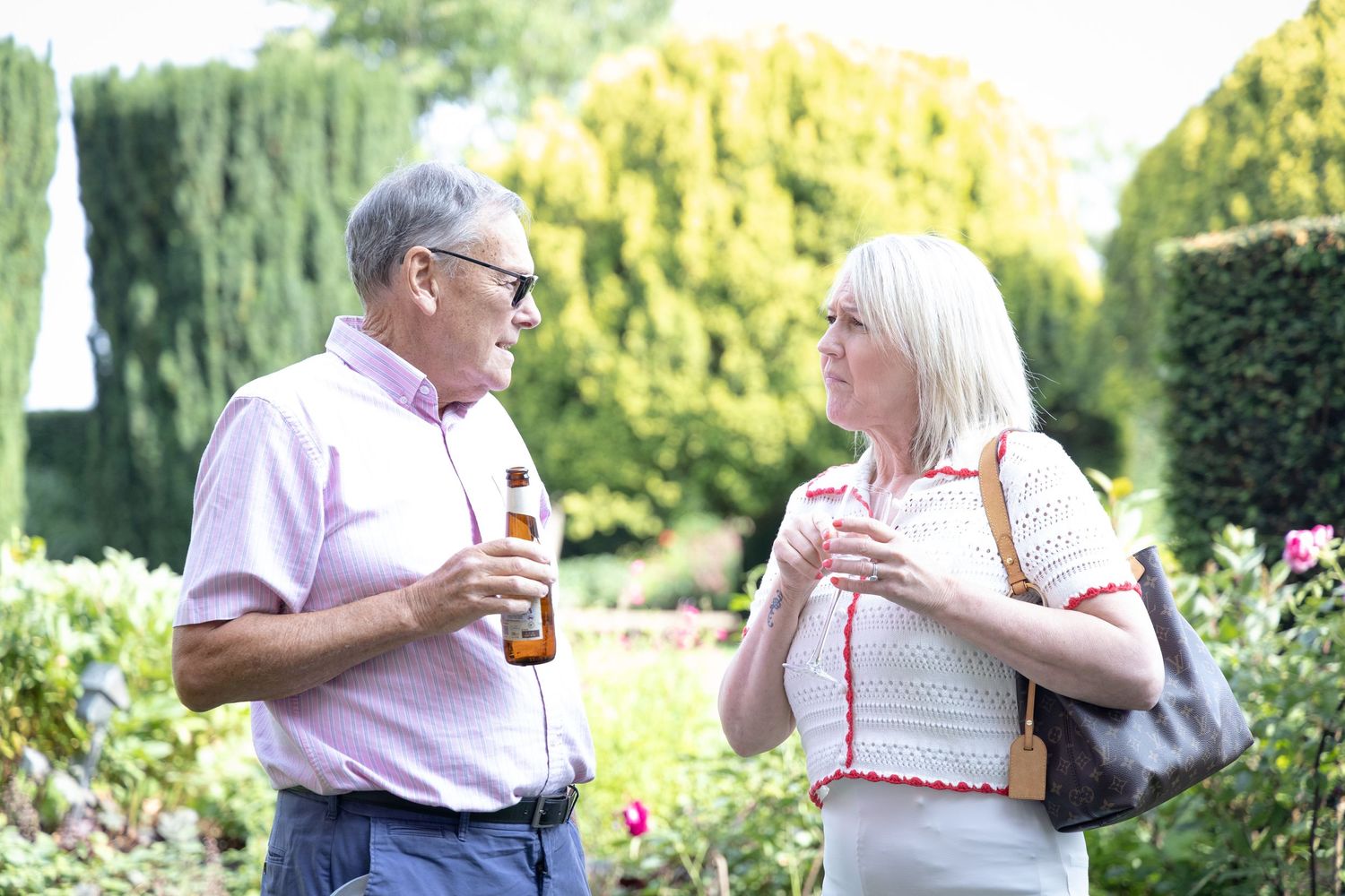 Two people enjoying drinks and conversation in a lush garden setting with trees and flowers.