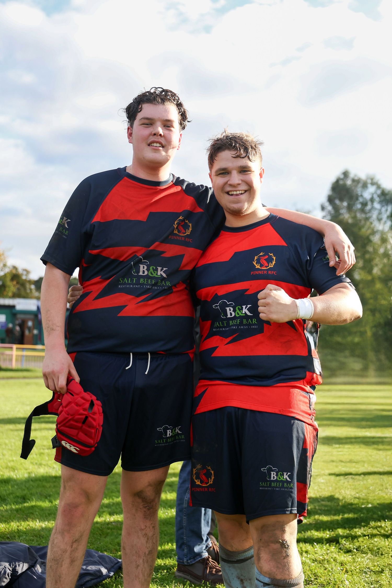 Two rugby players in red and black uniforms stand together smiling on a sunny field.
