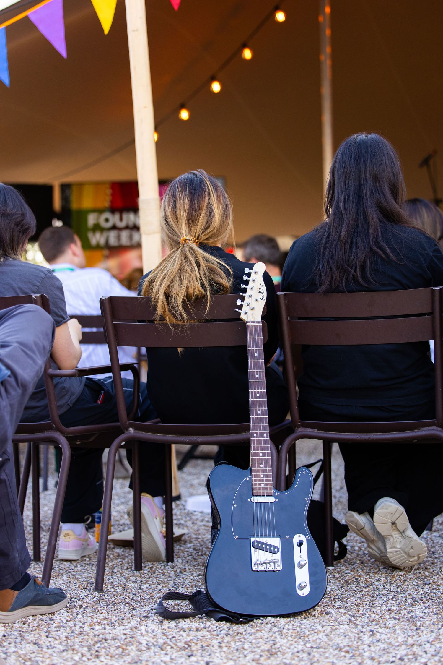 Electric guitar resting against wooden chairs at an outdoor event with colorful bunting and string lights.