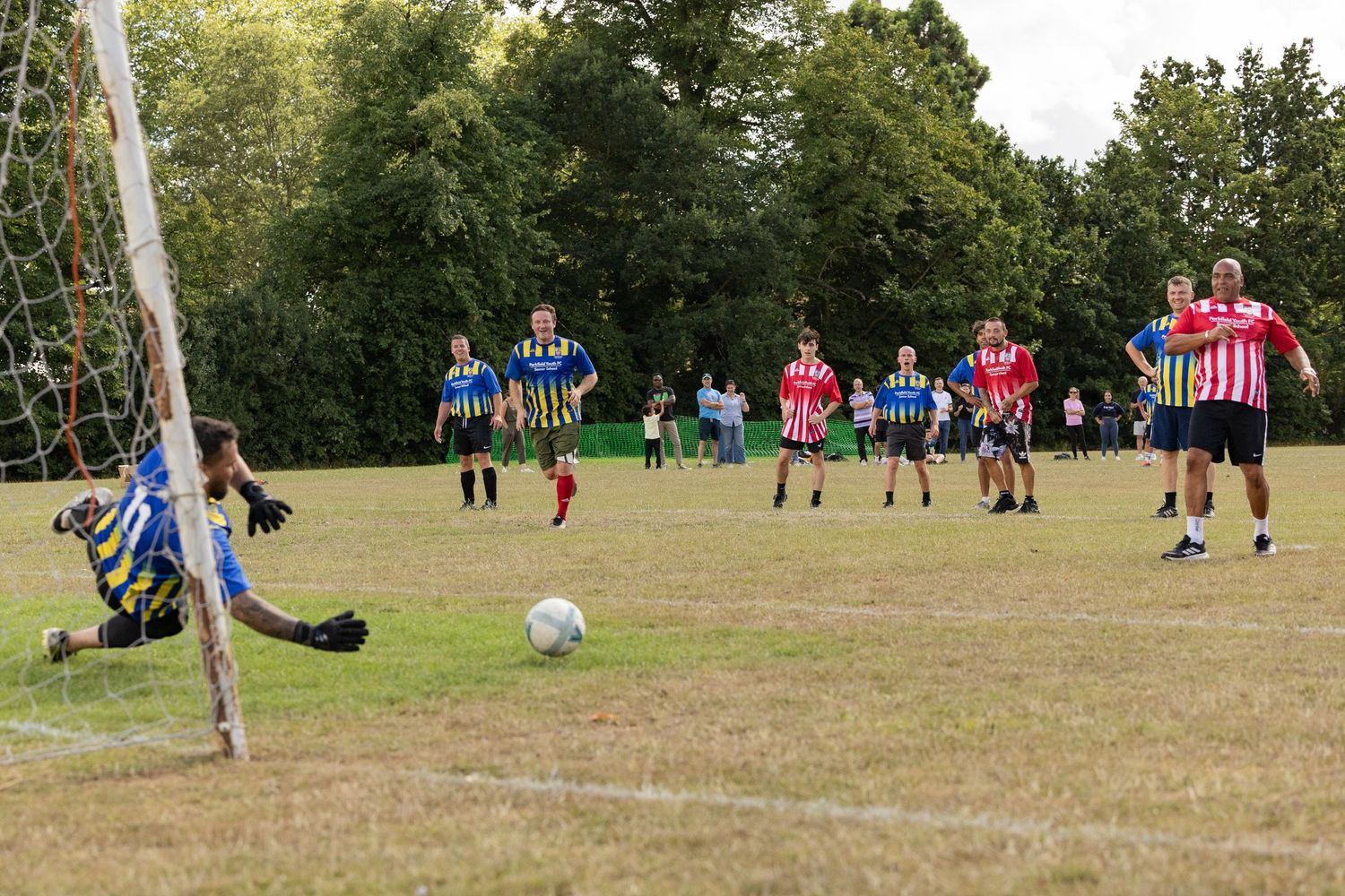 Players on a grass field during a soccer match as a goalie dives to block a shot at the goal.