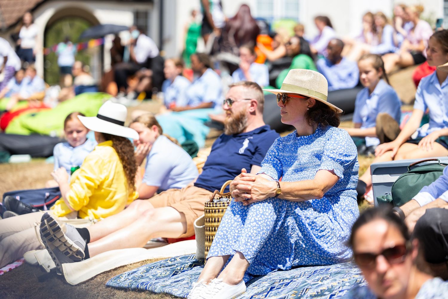 Crowd of spectators enjoying outdoor event while sitting on blankets and chairs in summer weather.
