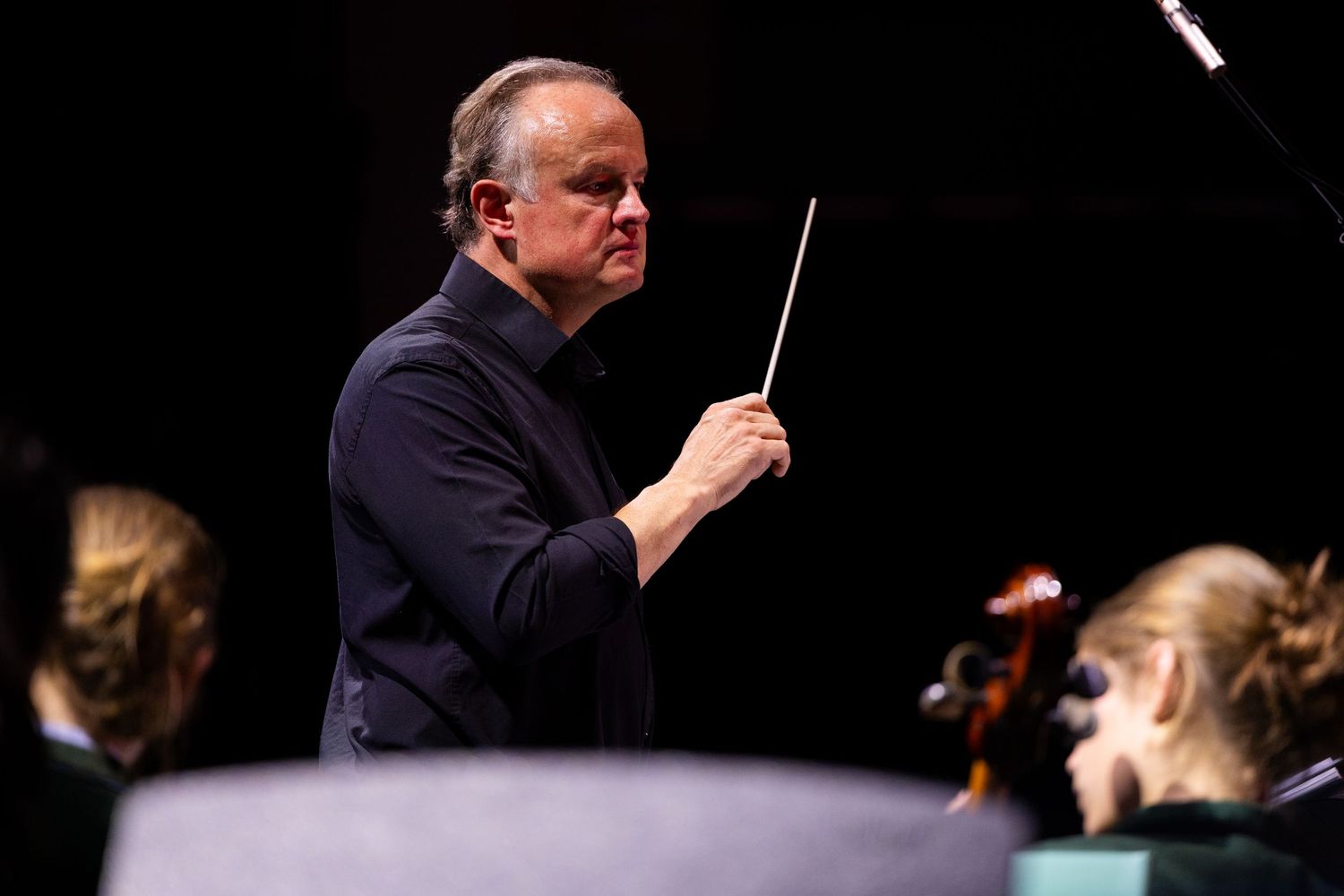 Orchestra conductor holds baton while directing musical performance on dark stage.
